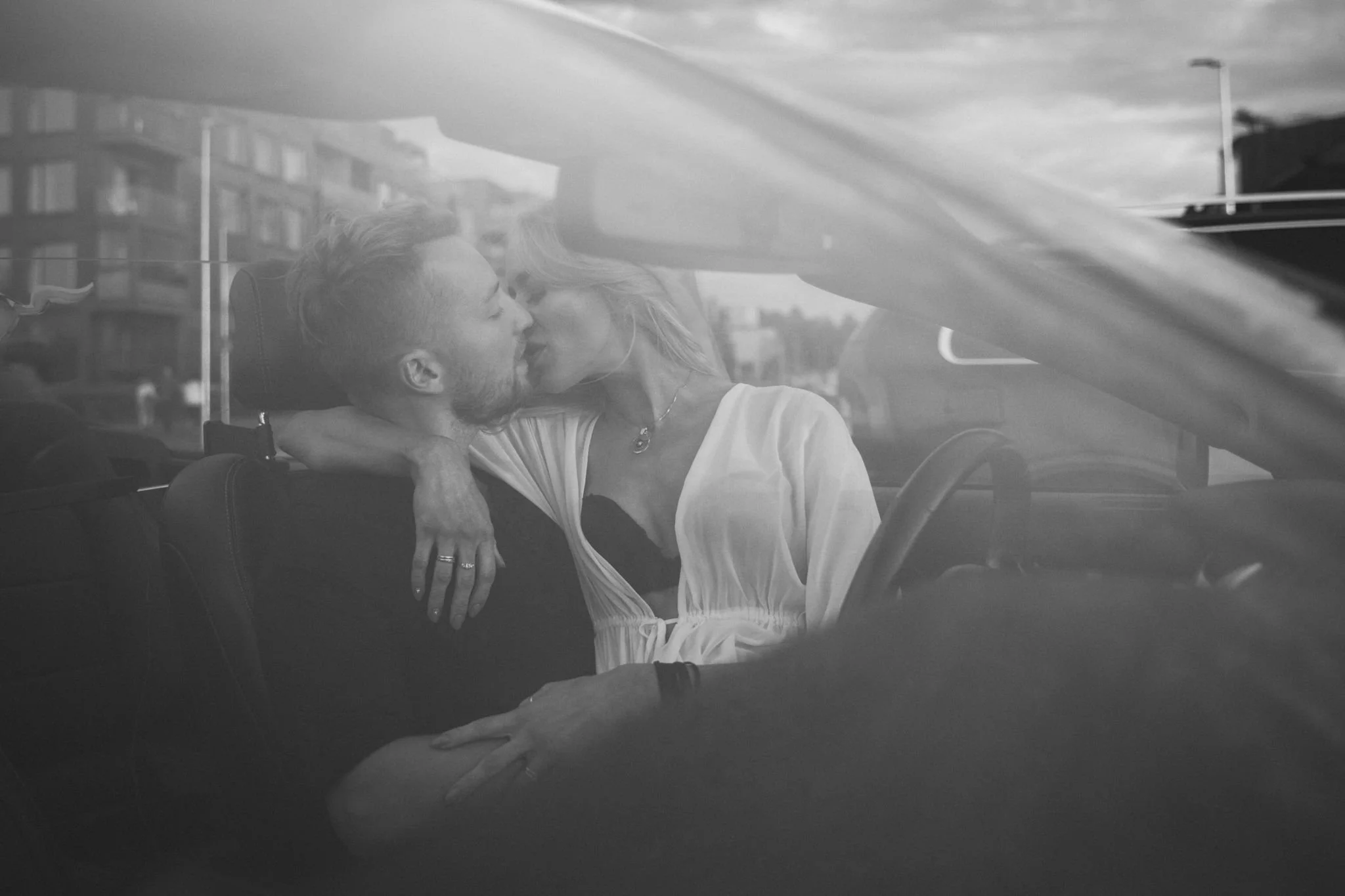 A couple sharing a kiss inside a car, with the reflection of buildings and the sky visible through the windshield.