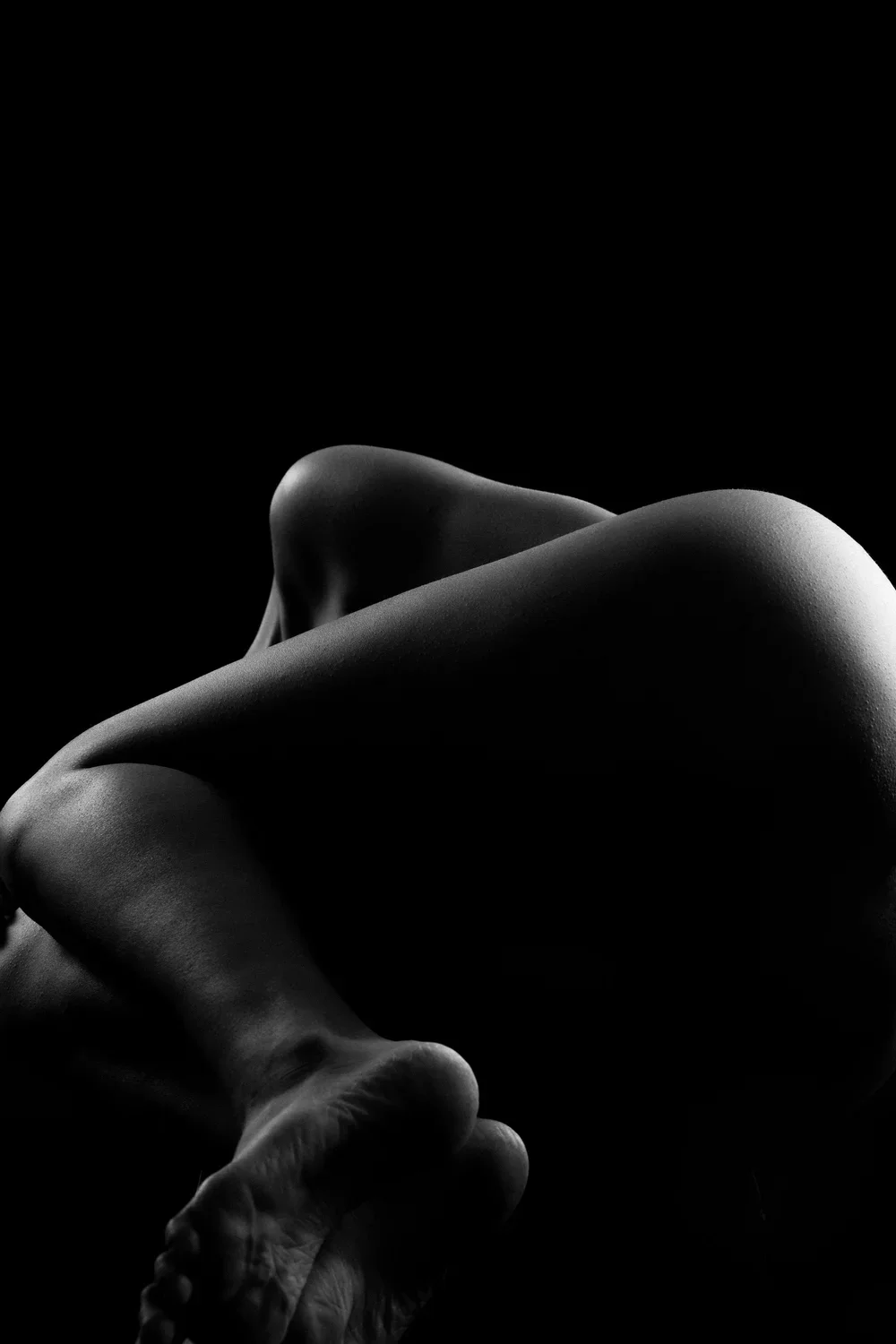 Black and white artistic photograph of a nude human figure in a curled position with dramatic lighting.