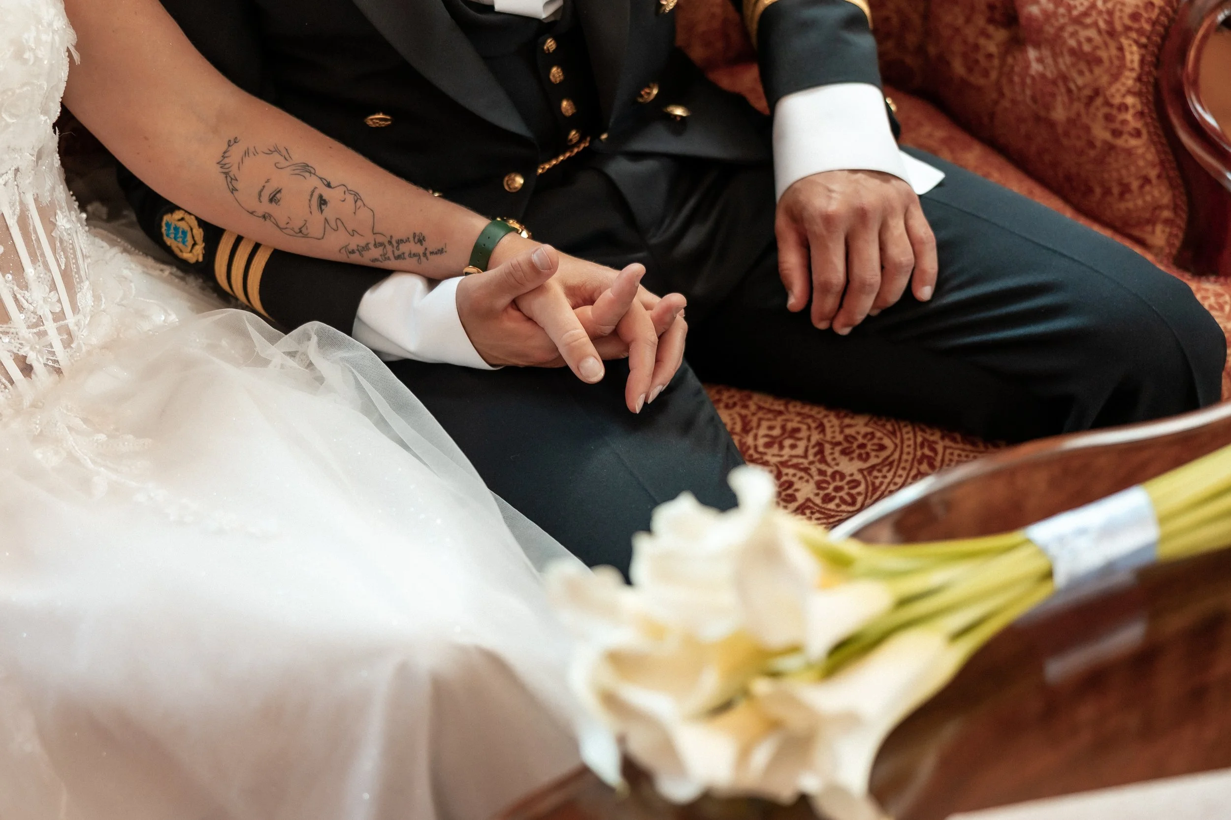 Close-up of a wedding couple sitting together, holding hands, with the bride's arm featuring a tattoo and the groom in a formal military uniform. The bride's dress and a bouquet are partially visible.