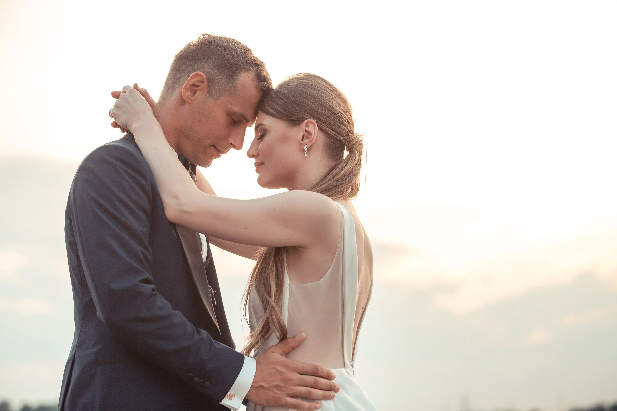 A couple dressed in formal attire embracing with their foreheads touching outdoors during sunset.