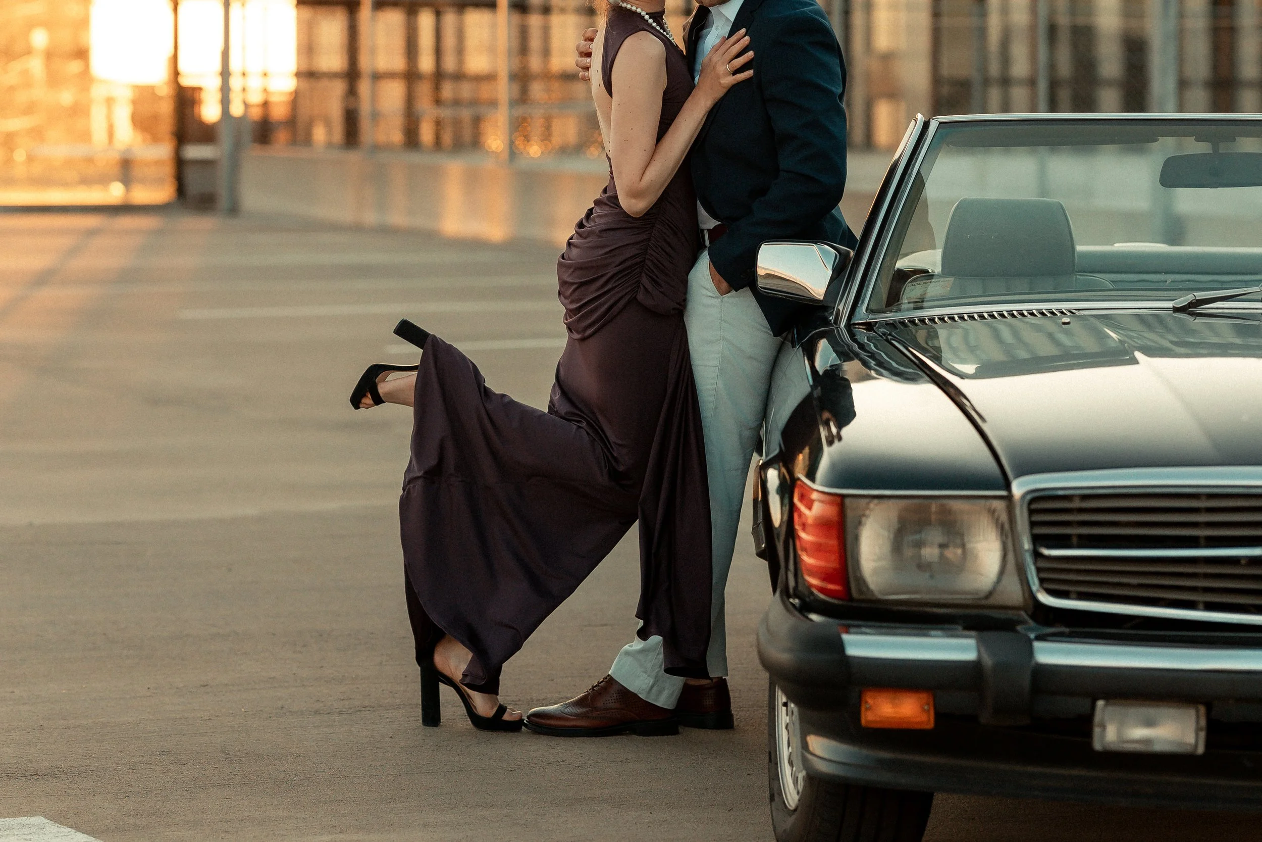 A couple dressed in formal attire sharing an intimate moment outdoors in the evening, with a vintage car parked nearby and city buildings in the background.