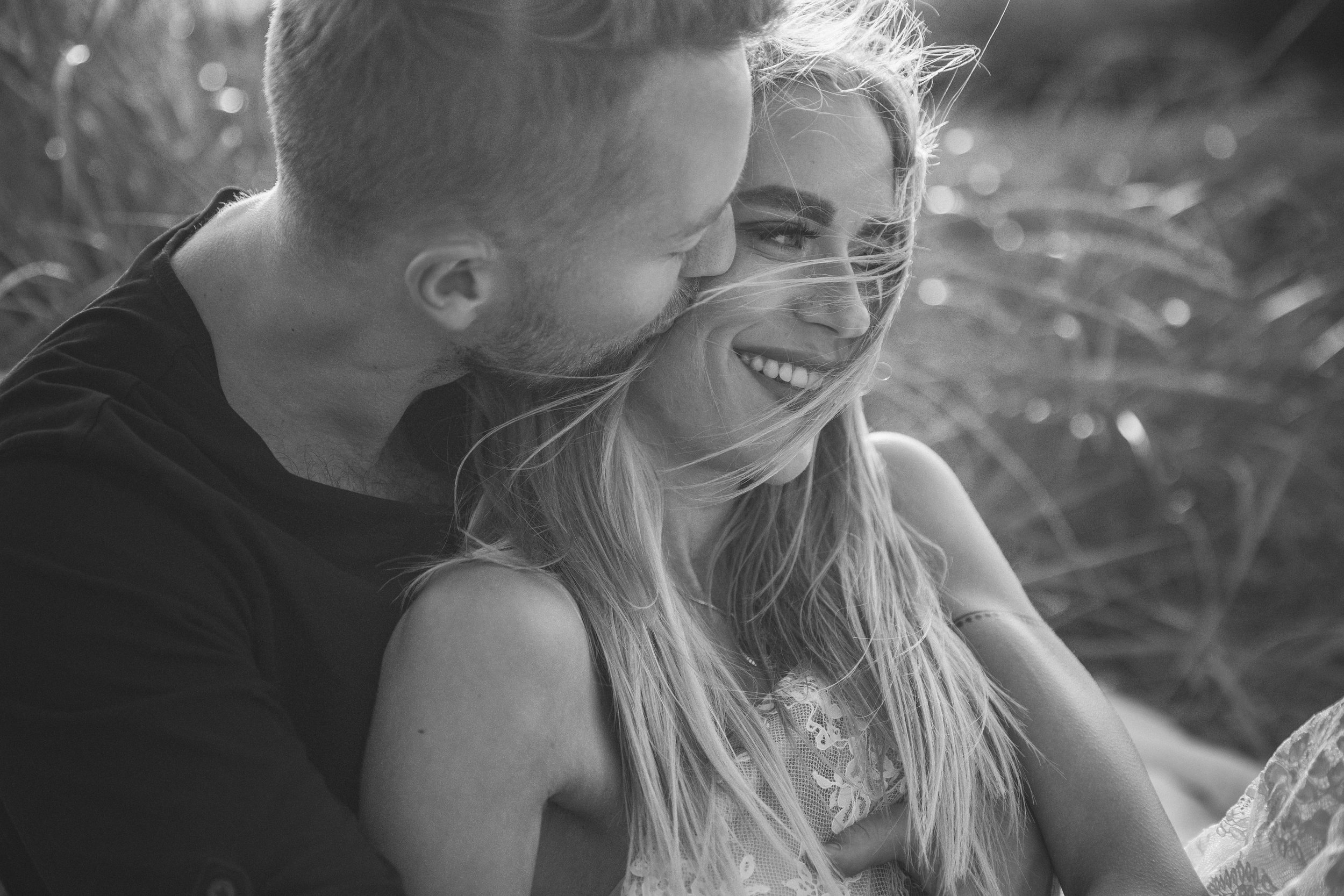 Black and white photo of a smiling blonde woman with wind-blown hair, close to a man with short hair and beard, outdoors near tall grass.