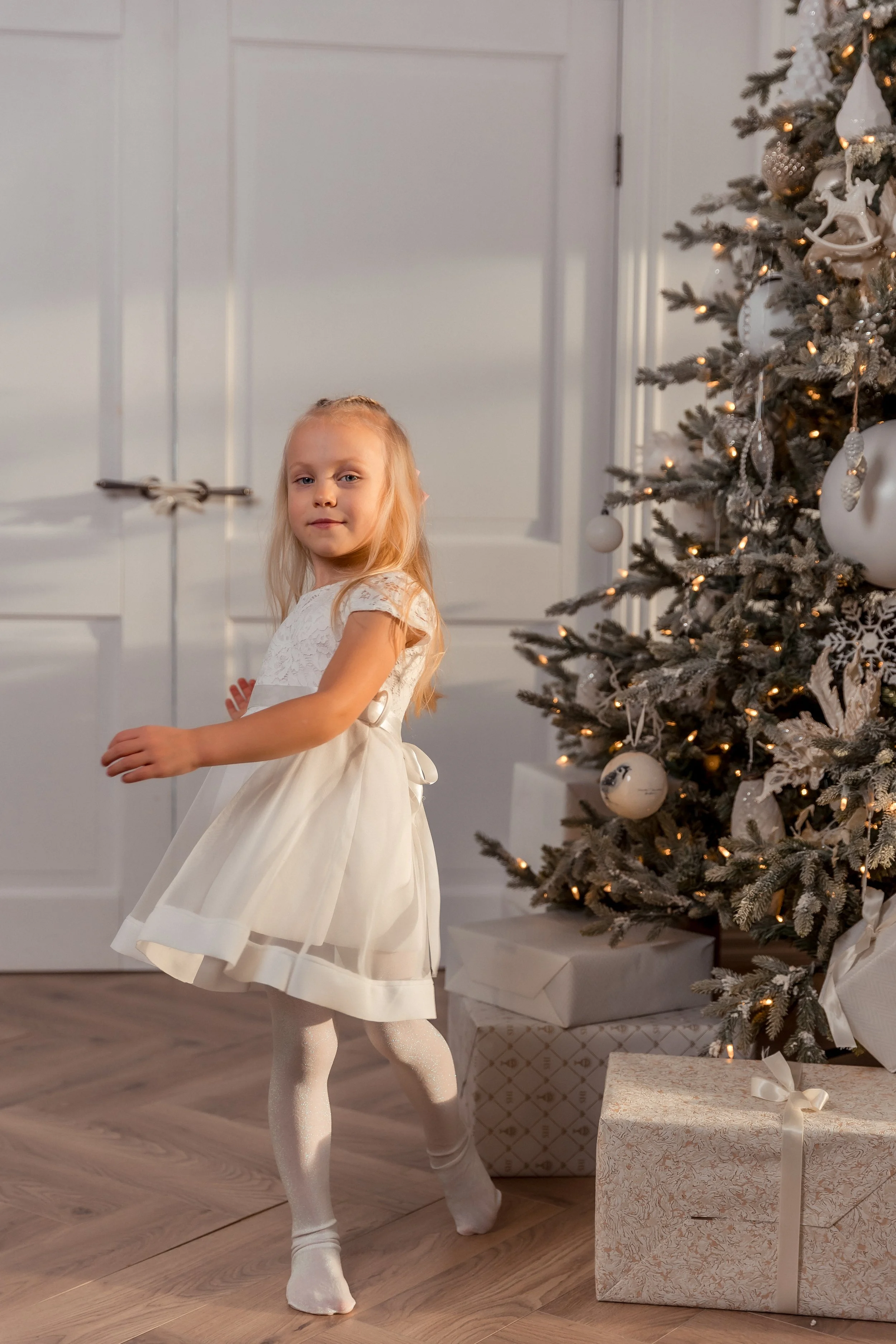A young girl wearing a white dress and tights is dancing near a decorated Christmas tree with lights and ornaments, with wrapped presents underneath.