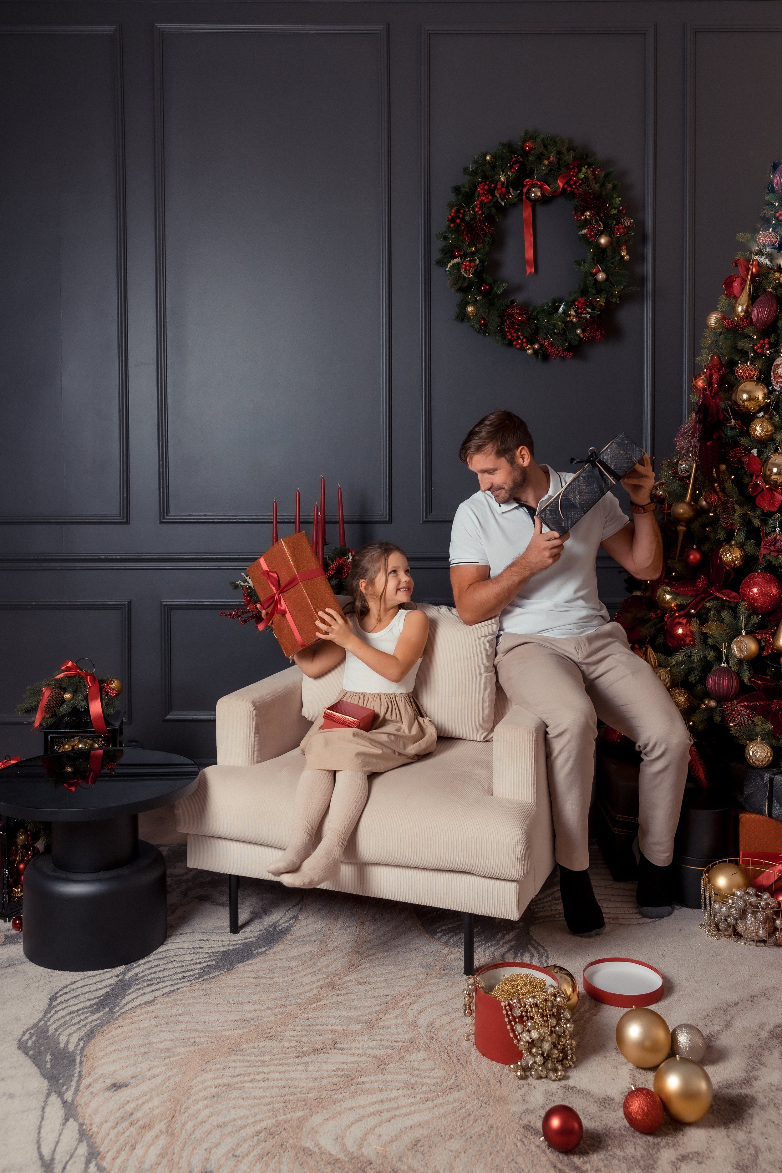 A man and a young girl sitting on a white couch in a living room decorated for Christmas. The girl is holding a large red gift box with a ribbon, and the man is holding a black gift box with a gold ribbon. Behind them, there's a decorated Christmas t