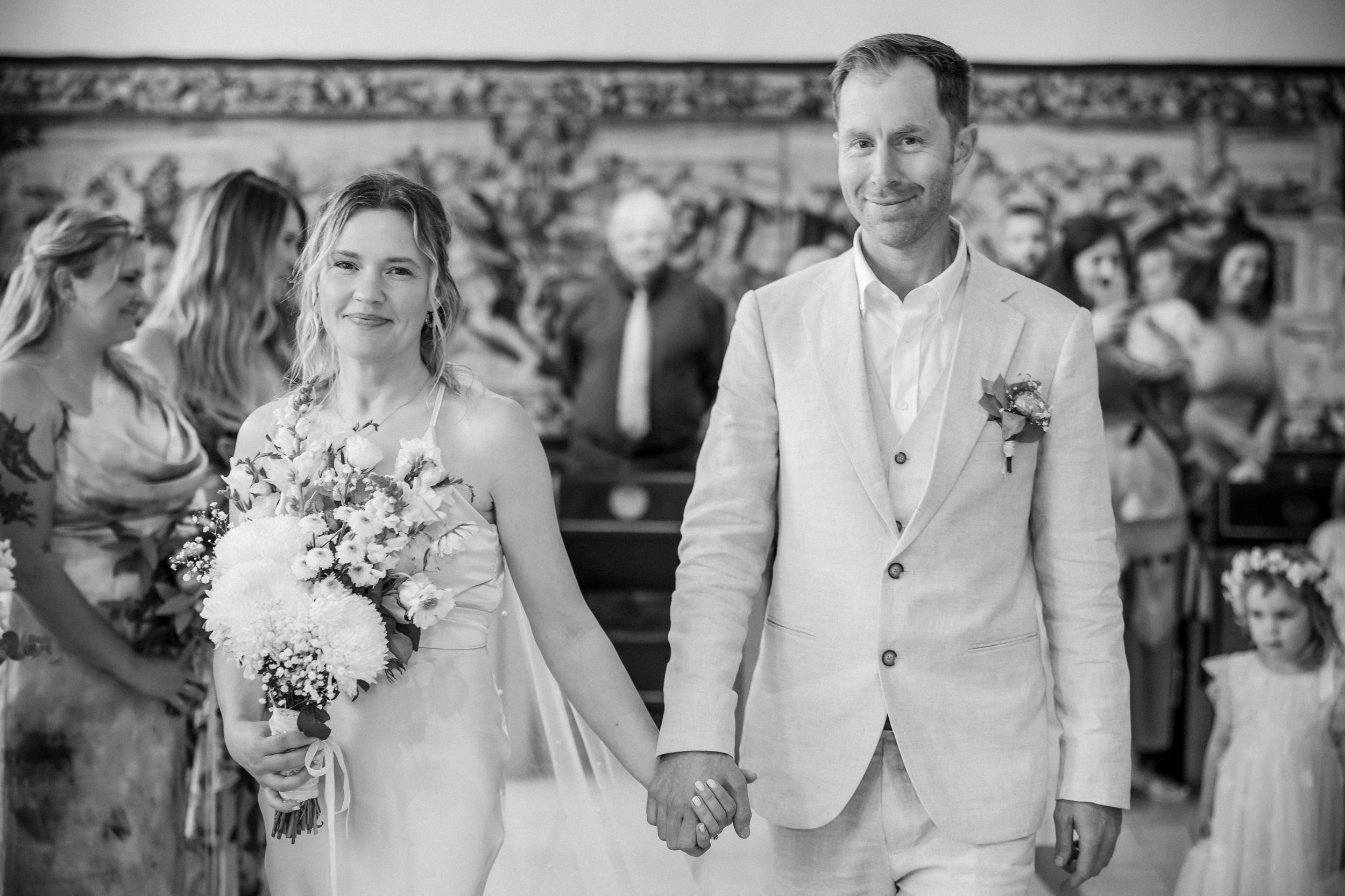 A black and white photo of a bride and groom holding hands during a wedding ceremony inside a church, with guests seated in the background.