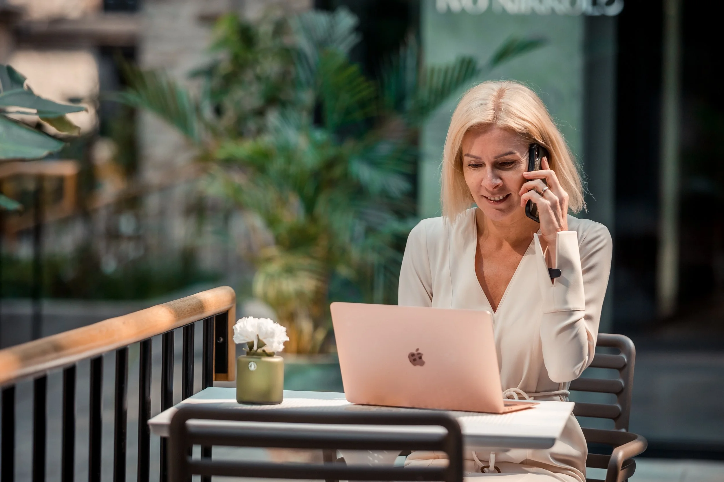 Woman with blonde hair sitting at a table in a cafe, talking on the phone, with a pink laptop and a small green vase with white flowers on the table, surrounded by green plants.