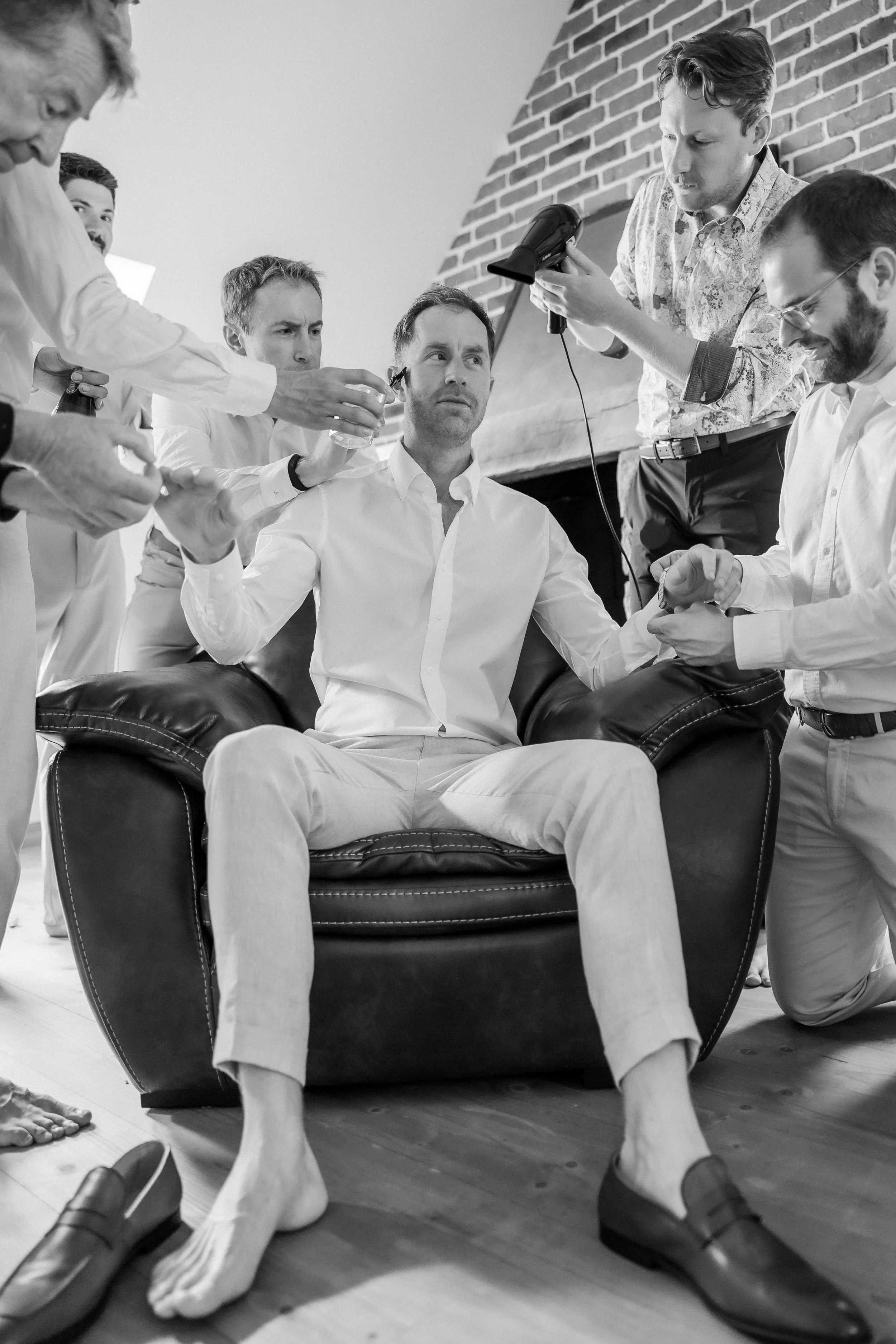 A black and white photo of a group of men in a room, with one man seated on a chair and the others standing around him. Some men are doing makeup or hairstyling, while others are holding equipment or looking at the seated man.