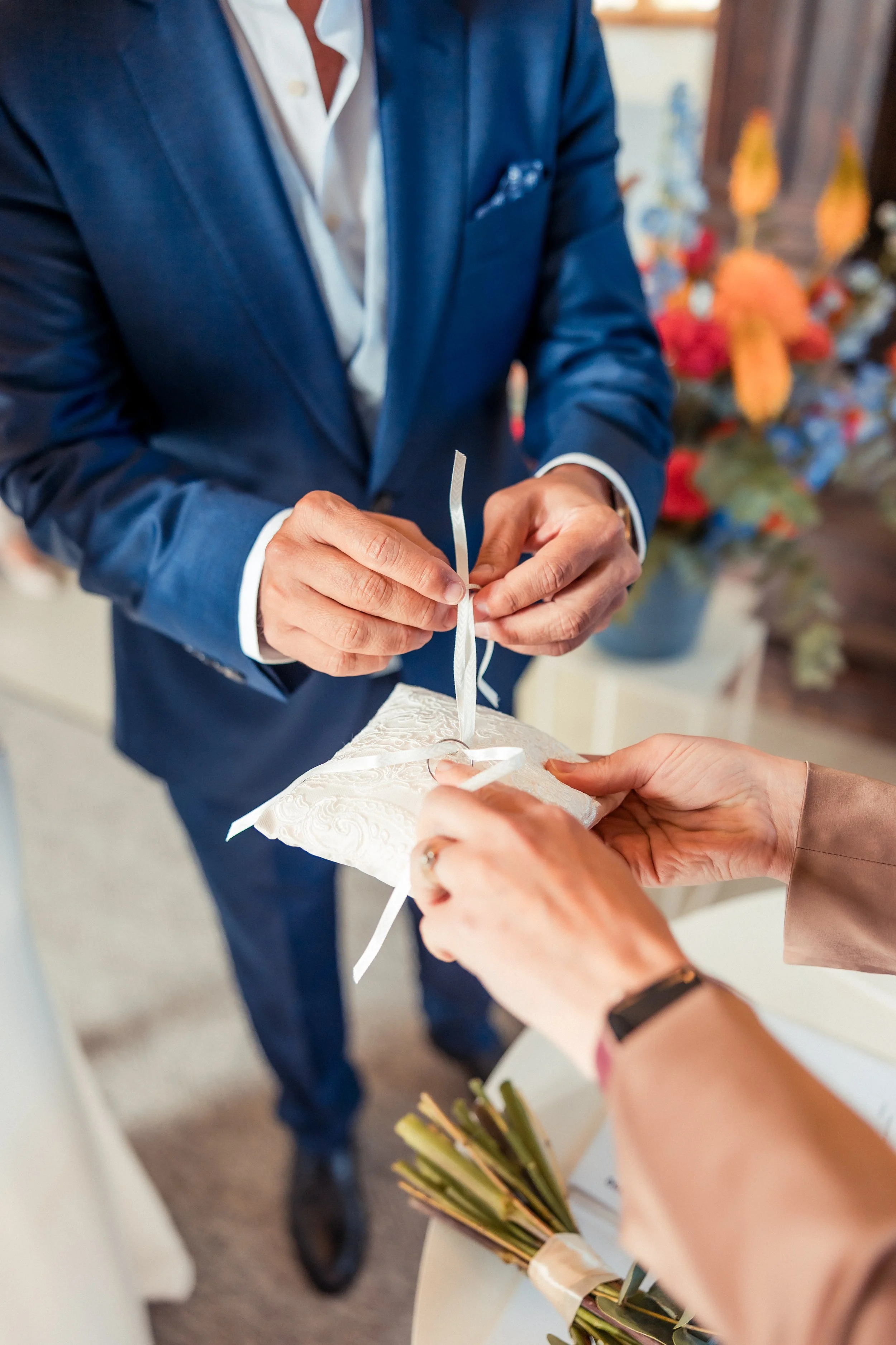 A man dressed in a blue suit is tying a white ribbon on a wedding cake. Two women are holding a bouquet and helping with the ribbon. A bouquet of flowers is on the table in the foreground.