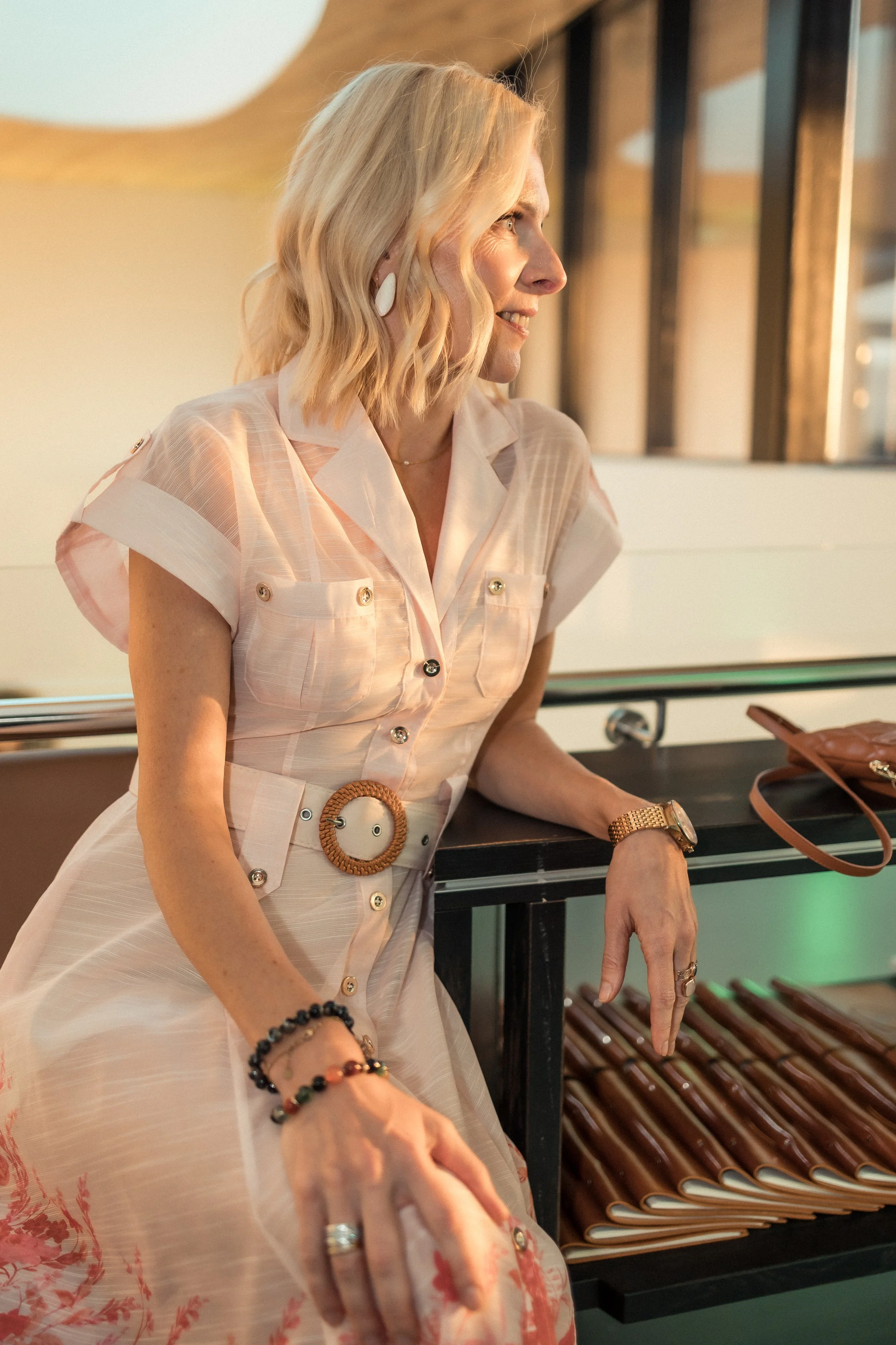 A woman with blonde hair wearing a light-colored, patterned dress with buttons and a wide belt, leaning against a railing near a display of rolled-up mats or designs, with large window panes in the background.