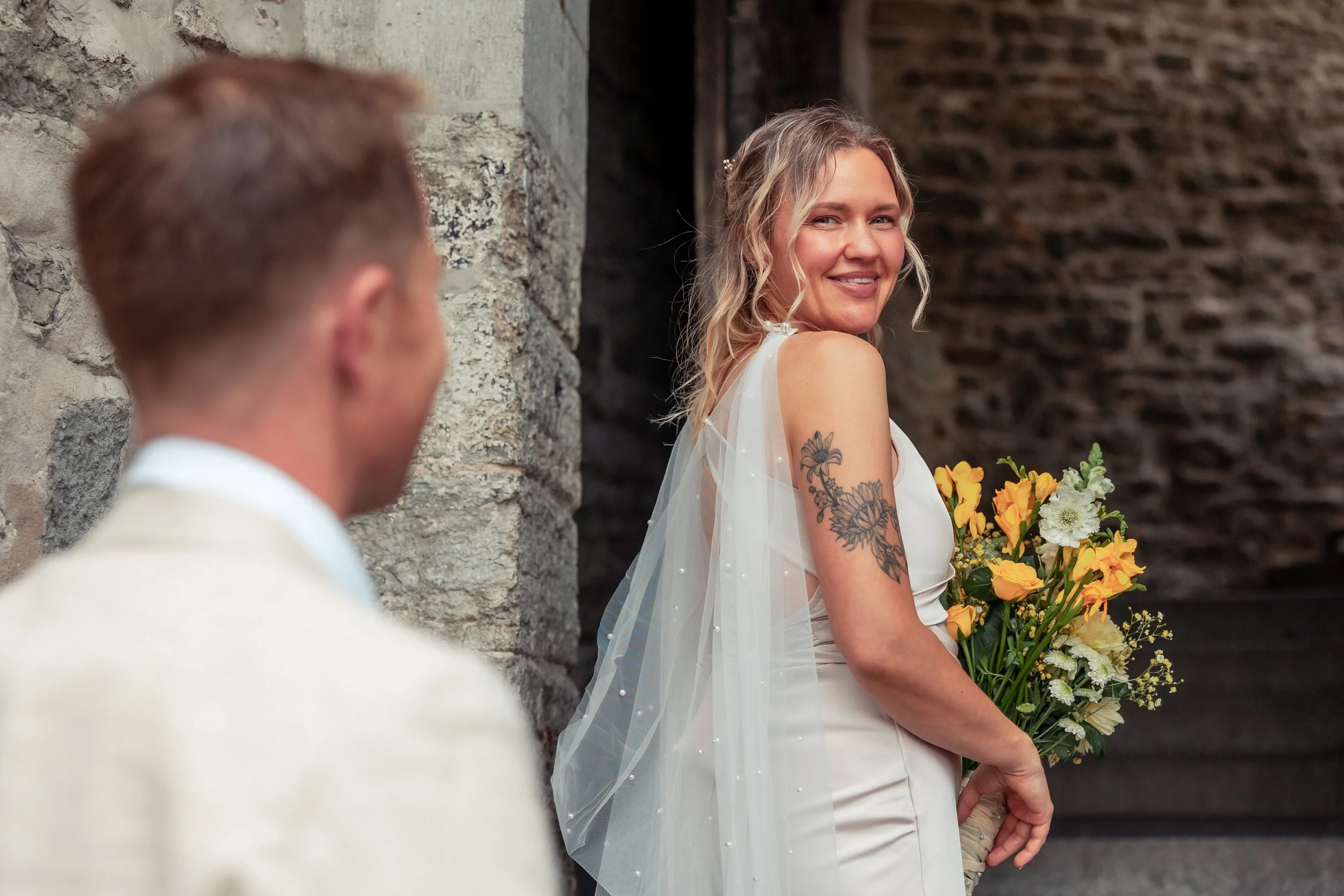 A woman in a white dress with yellow and white flowers in her hands, looking at a man in a white suit, with a brick wall and stone wall background at what appears to be a wedding or special celebration.