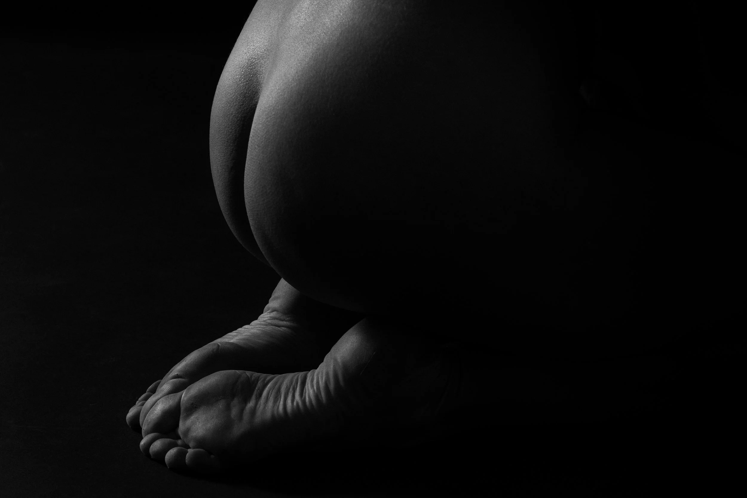 Black and white photo of a person kneeling with hands clasped on the floor, their back turned to the camera, in a dark setting.