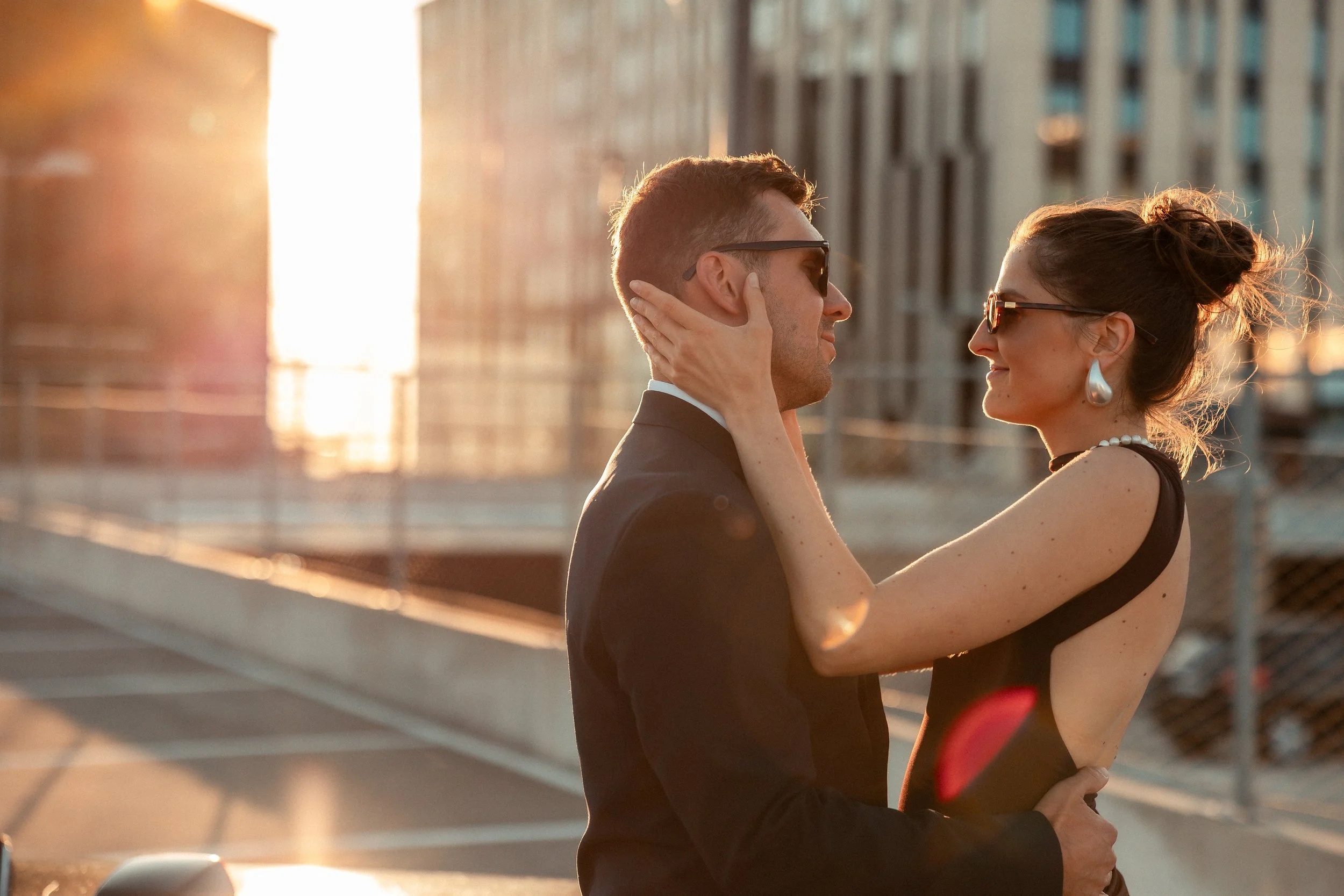 A couple is embracing each other while smiling, wearing sunglasses, on a rooftop during sunset in the city.