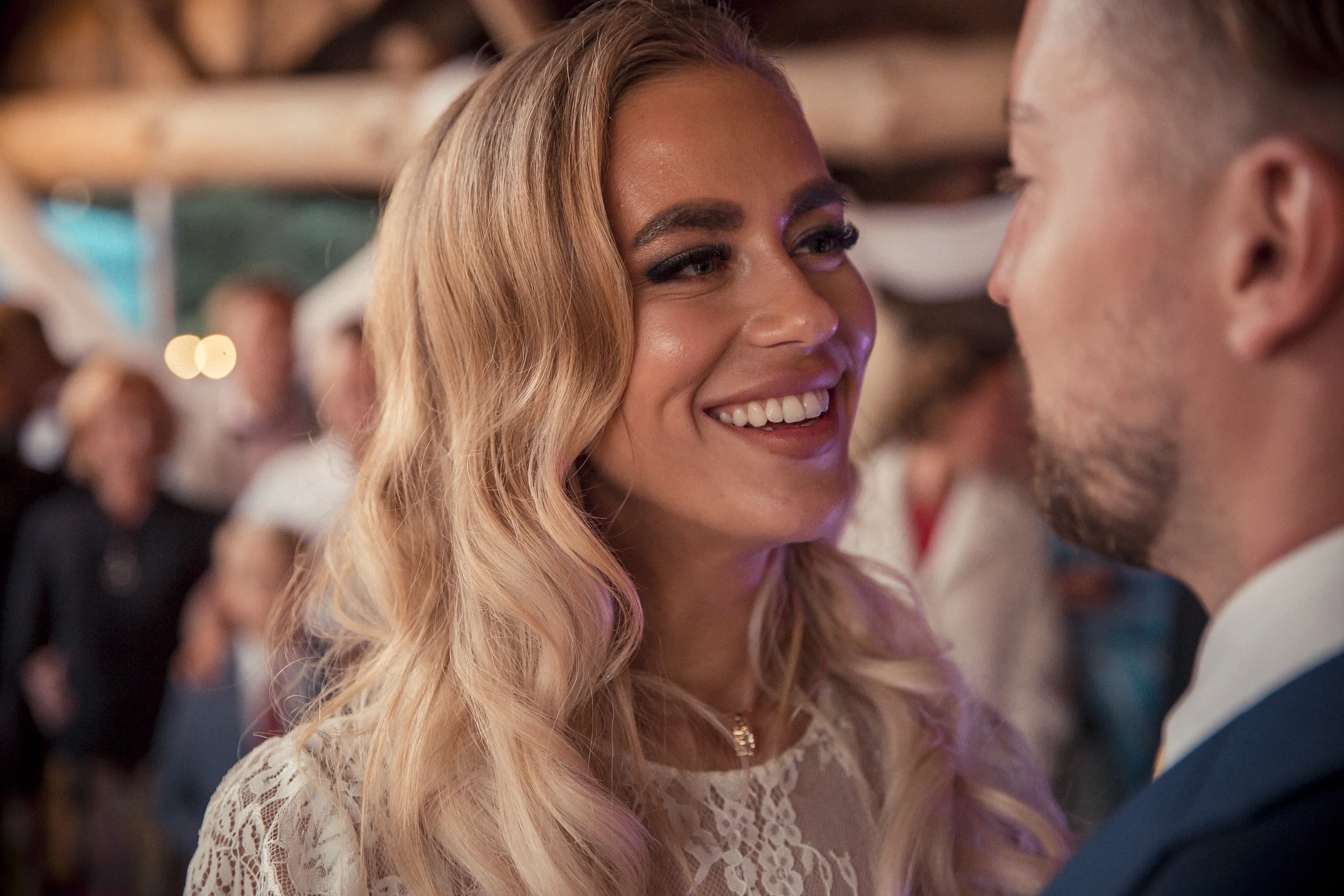 A smiling woman with long, wavy blonde hair looking at a man at a social event, background blurred with other people.