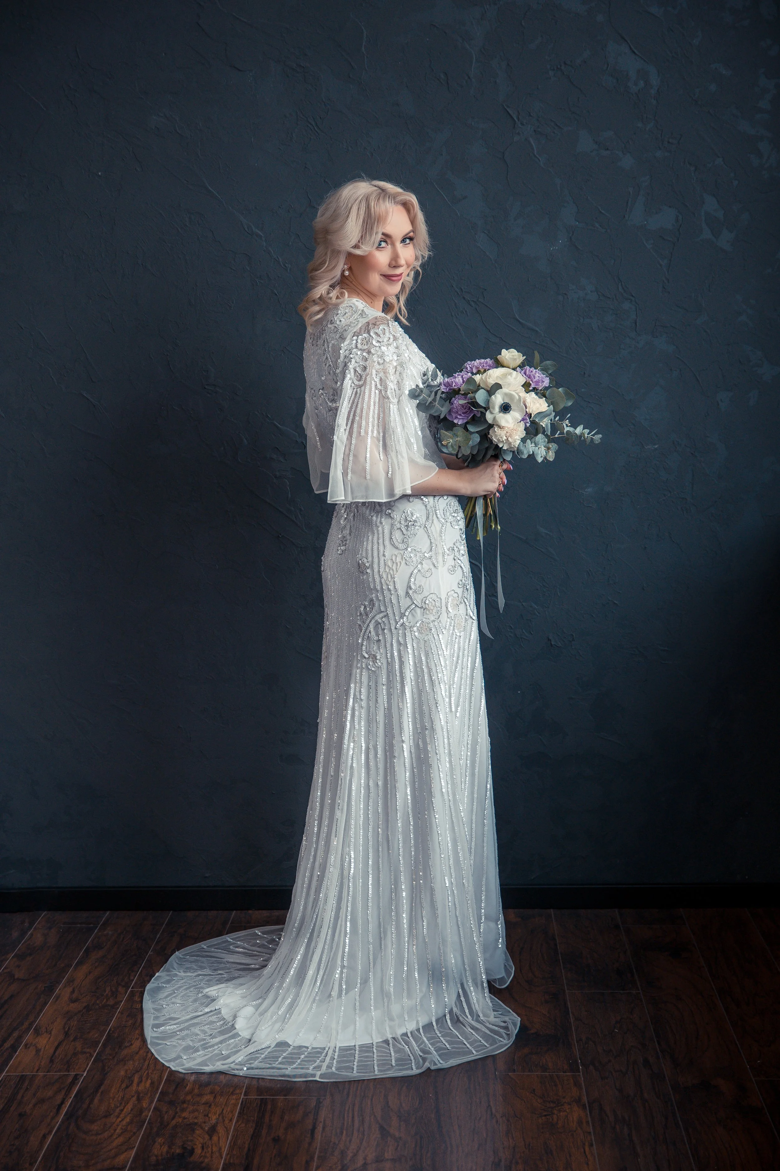 A woman in a white, beaded, and sequined wedding dress holding a bouquet of white and purple flowers stands against a dark gray textured wall. She has wavy blonde hair and is smiling at the camera.