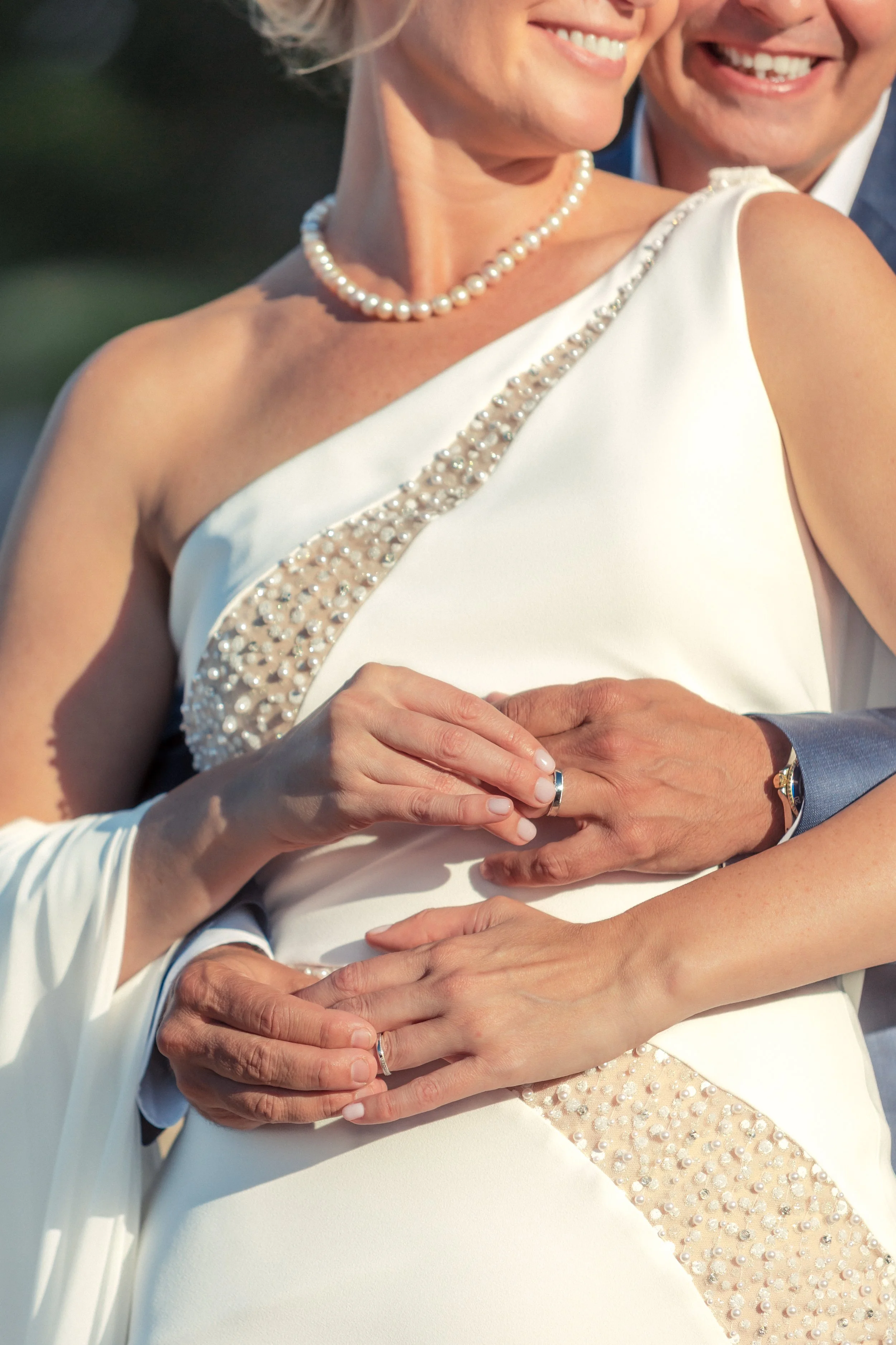 Close-up of a wedding couple with their hands intertwined, both wearing wedding bands. The bride is smiling, wearing a pearl necklace, and a white gown with pearl embellishments. The groom's face is partially visible, smiling, wearing a blue suit. Th