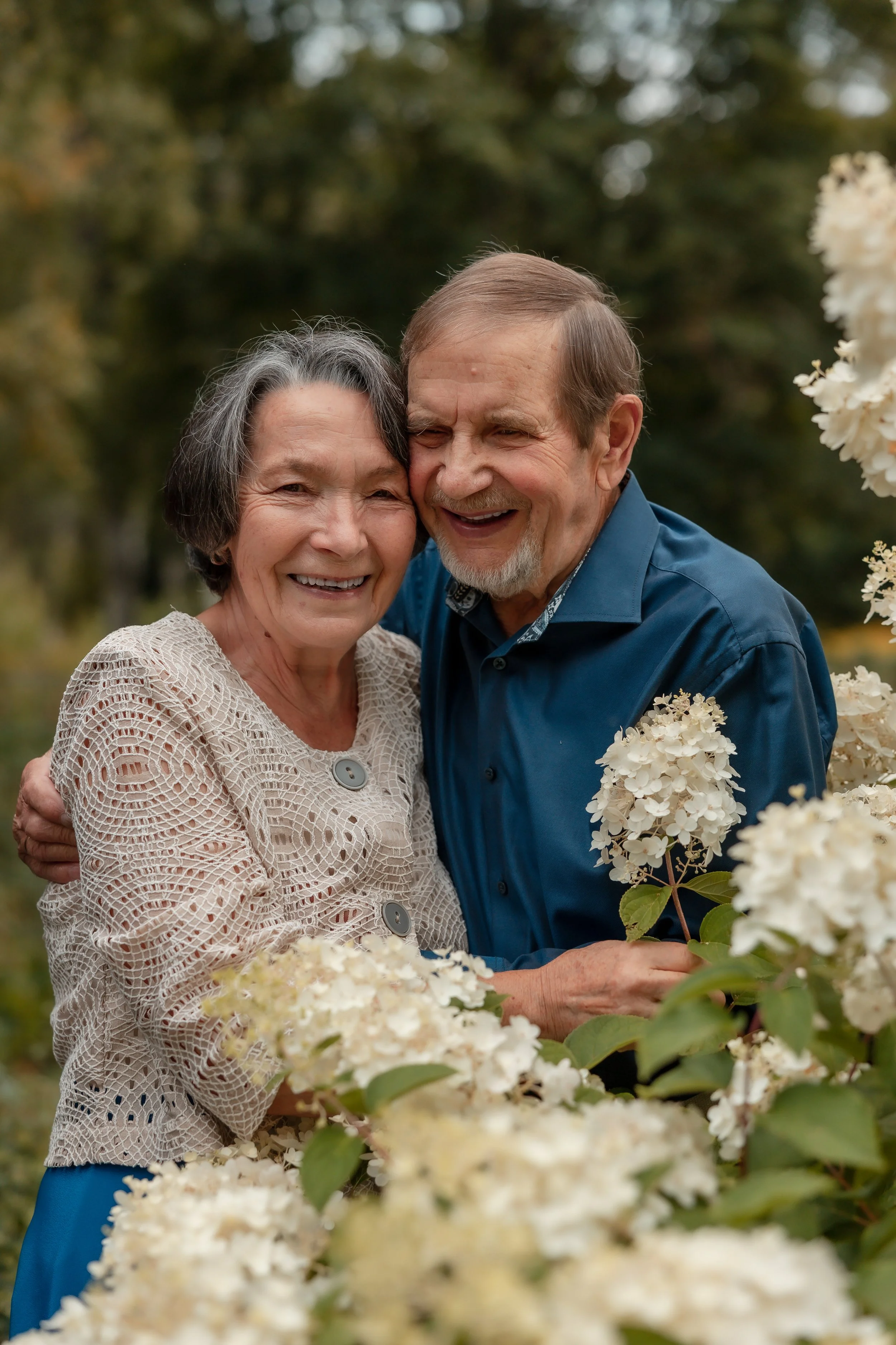 An elderly couple smiling and hugging in a garden with white flowering bushes, surrounded by greenery.