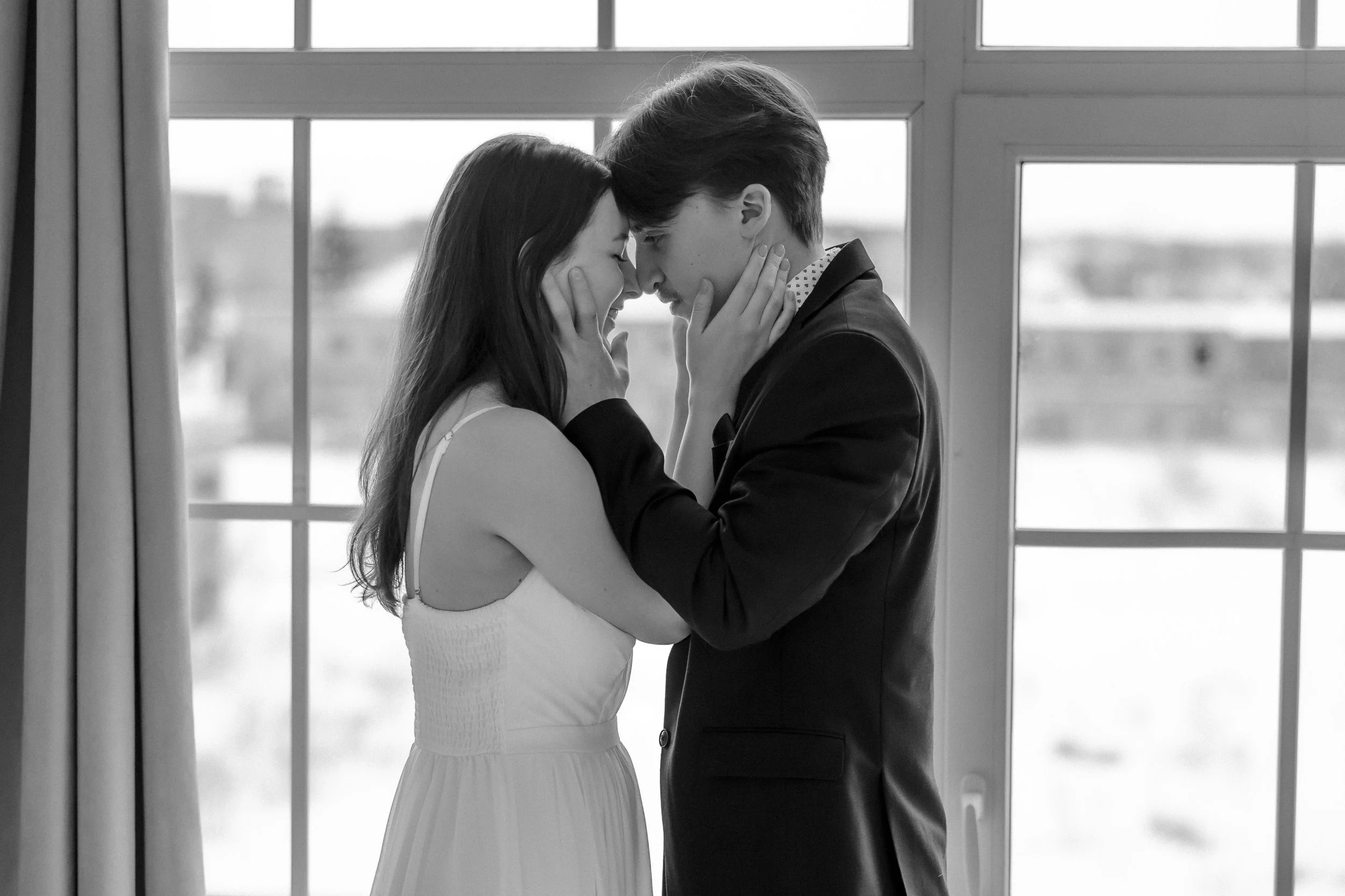 Black and white photo of a couple, a woman in a white dress and a man in a suit, standing close inside, touching foreheads, with their eyes closed, in front of large window with curtains.