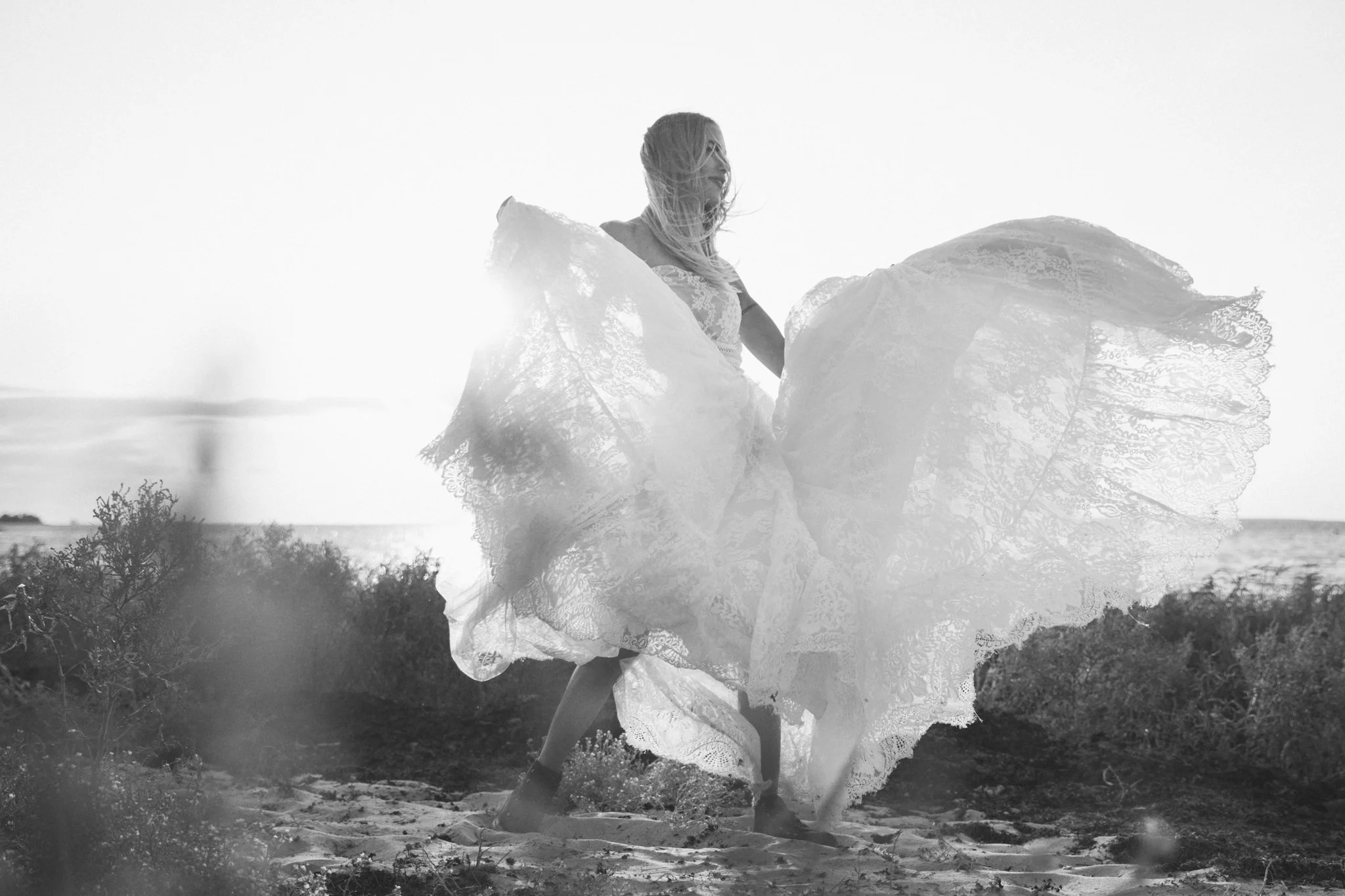 A black-and-white photo of a woman wearing a lace dress, standing outdoors with her dress flowing in the wind. She appears to be smiling and enjoying the moment, with a bright sky behind her and some bushes and trees at her feet.