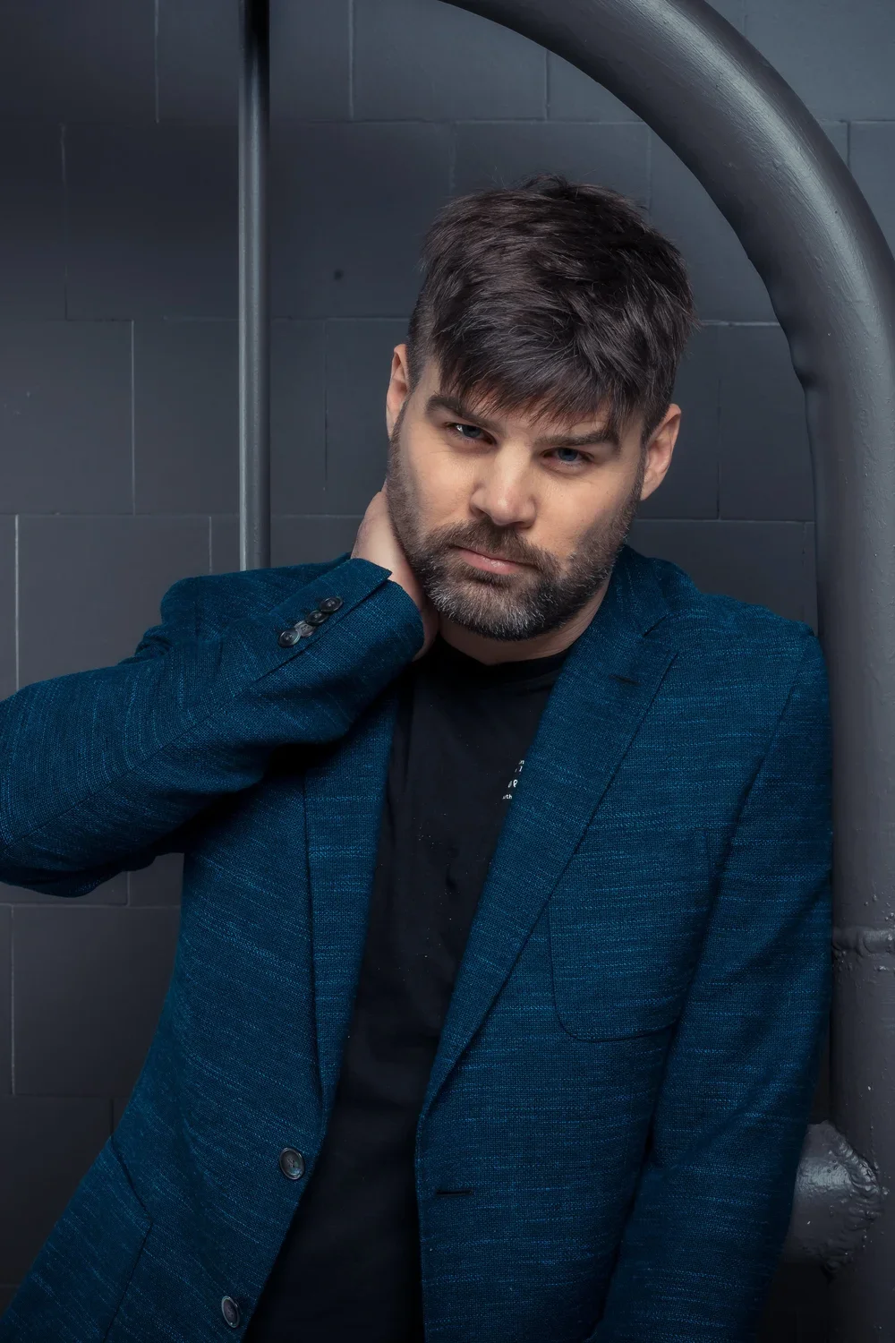 A man with dark hair and beard wearing a blue blazer, standing against a gray wall with pipes, posing with his hand on his neck and looking at the camera.