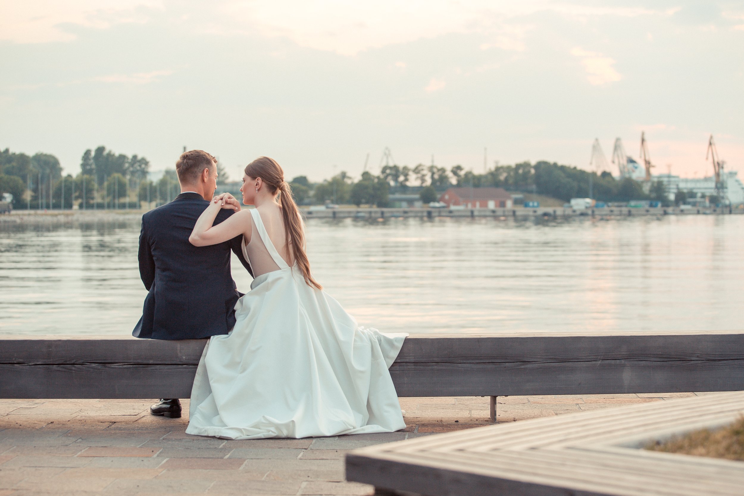 A bride and groom sitting on a bench by the water, facing each other, with the bride in a white wedding dress and the groom in a black suit, during sunset or late afternoon.
