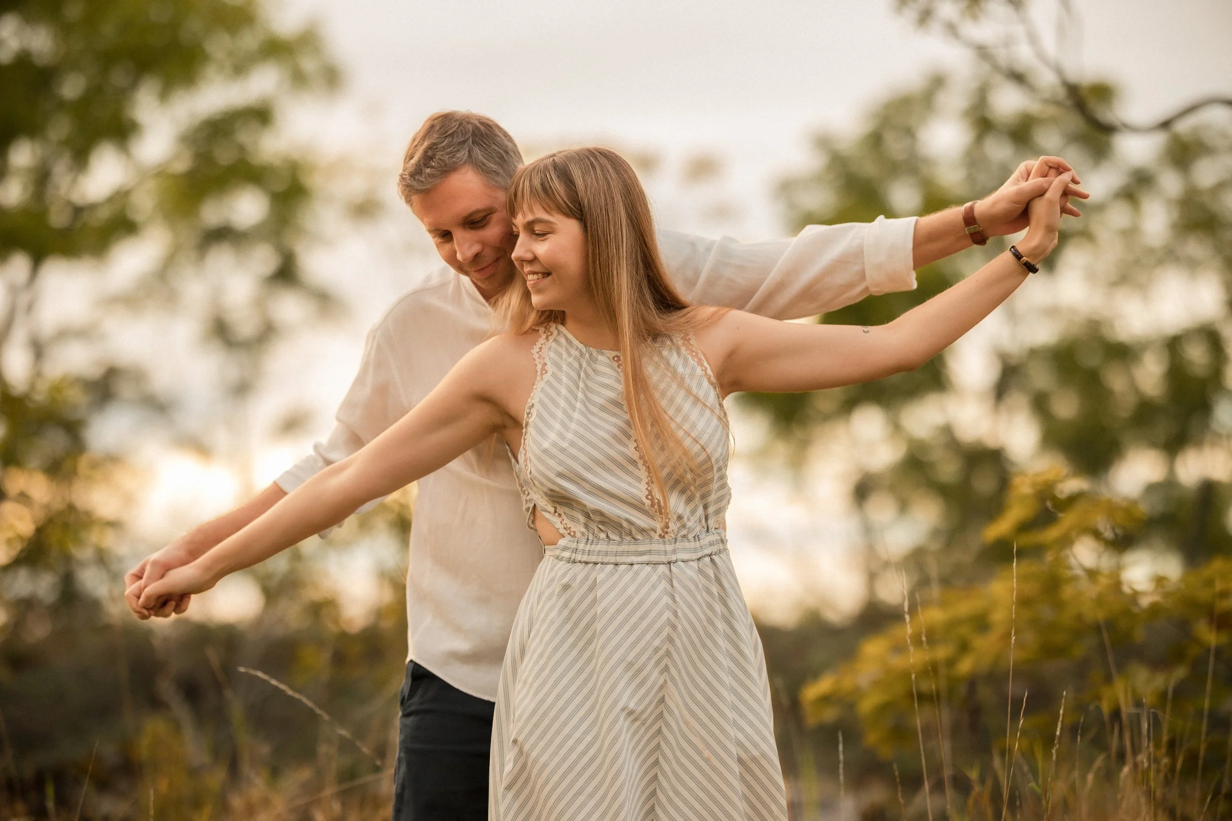 A young couple standing outdoors, holding hands and leaning their heads together, with the woman extending her arms out to the sides. They are smiling and appear to be enjoying a moment together in a natural setting with trees in the background.