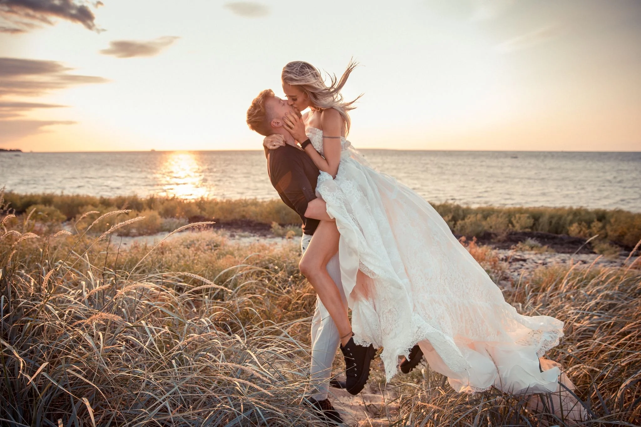 A couple sharing a romantic kiss on a beach at sunset, with the woman in a white dress and the man in dark clothing, surrounded by tall grass.