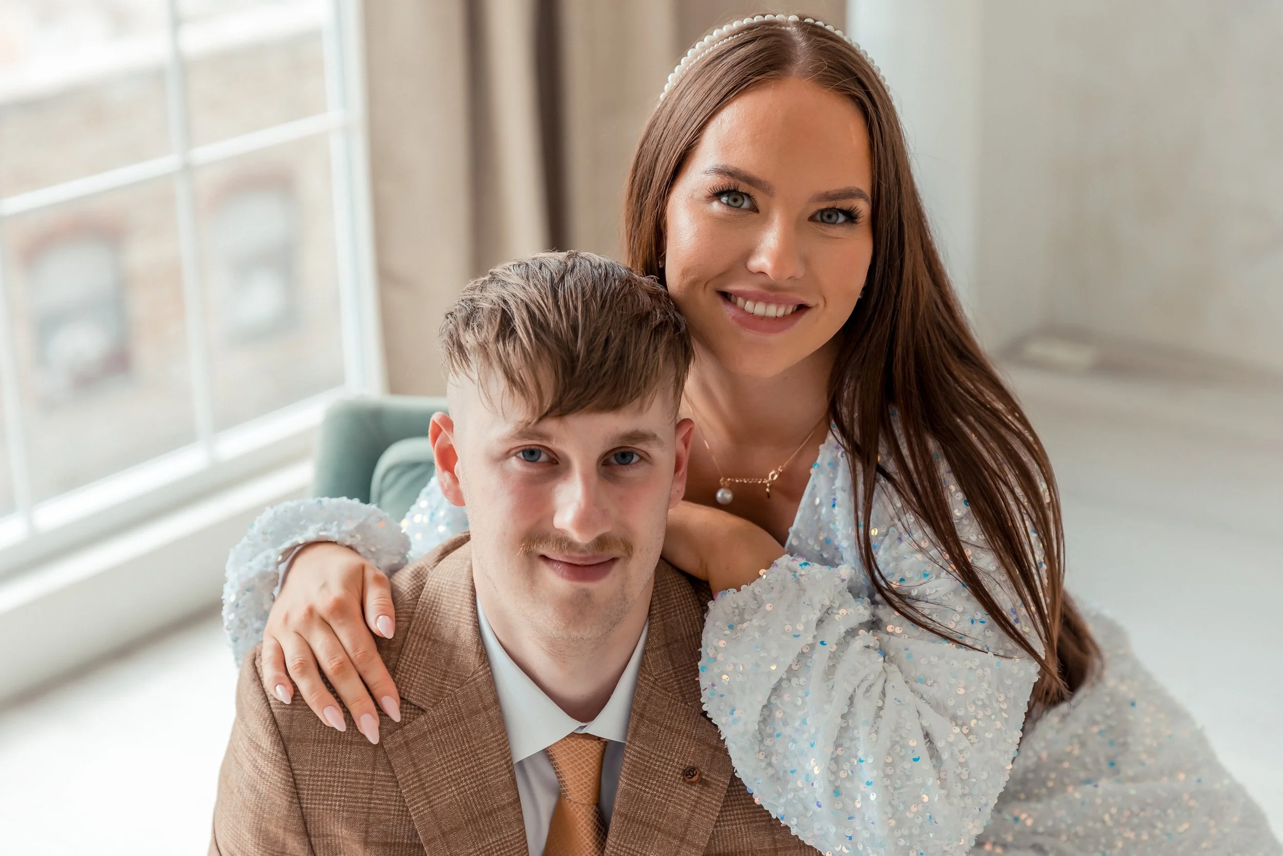 A smiling woman with long reddish-brown hair and a pearl headband embraces a young man with light brown hair and blue eyes, both dressed in formal attire, indoors with natural light.