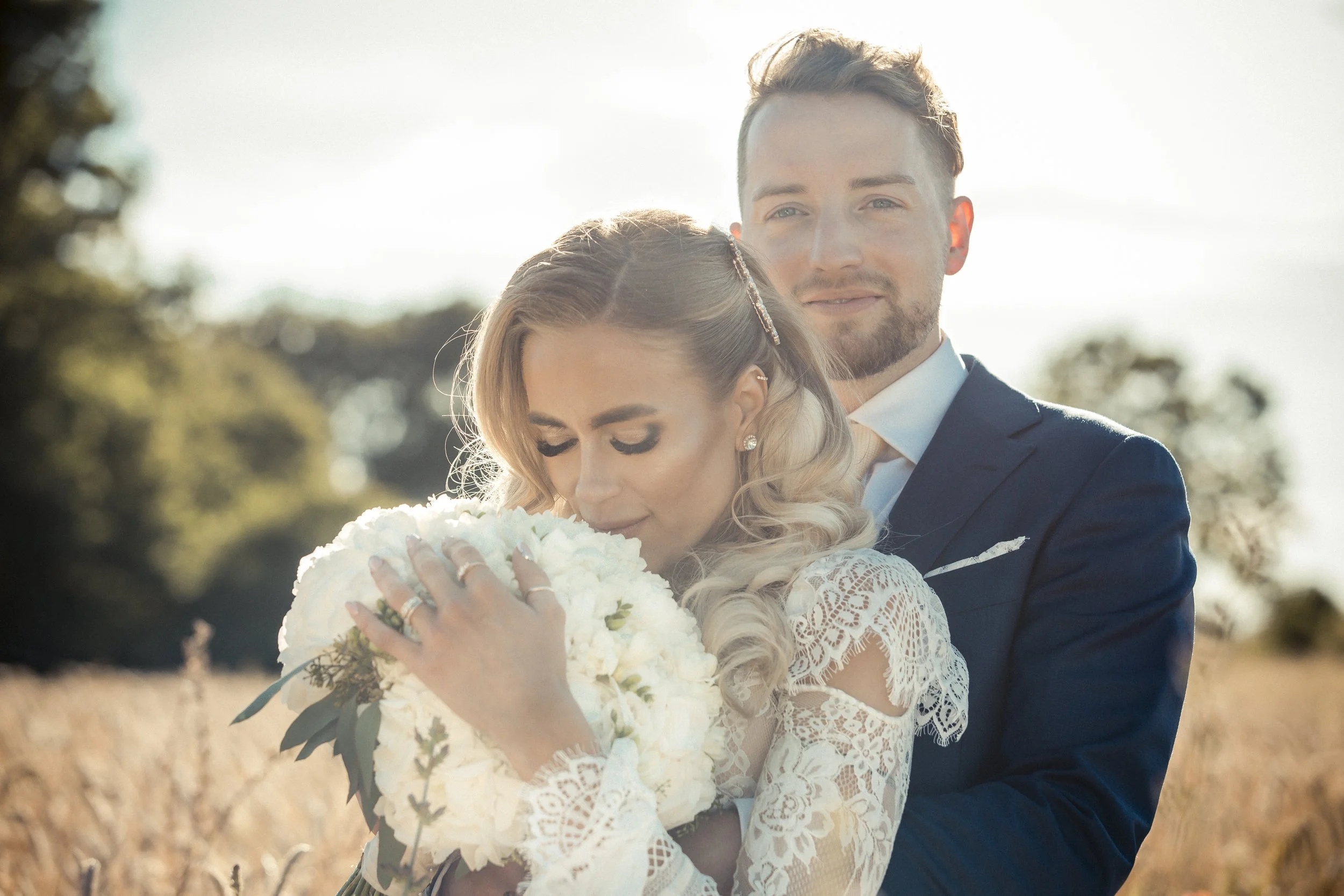 A newlywed couple embracing outdoors during sunset, with the woman holding a bouquet of white flowers and wearing a lace wedding dress and the man in a dark suit.