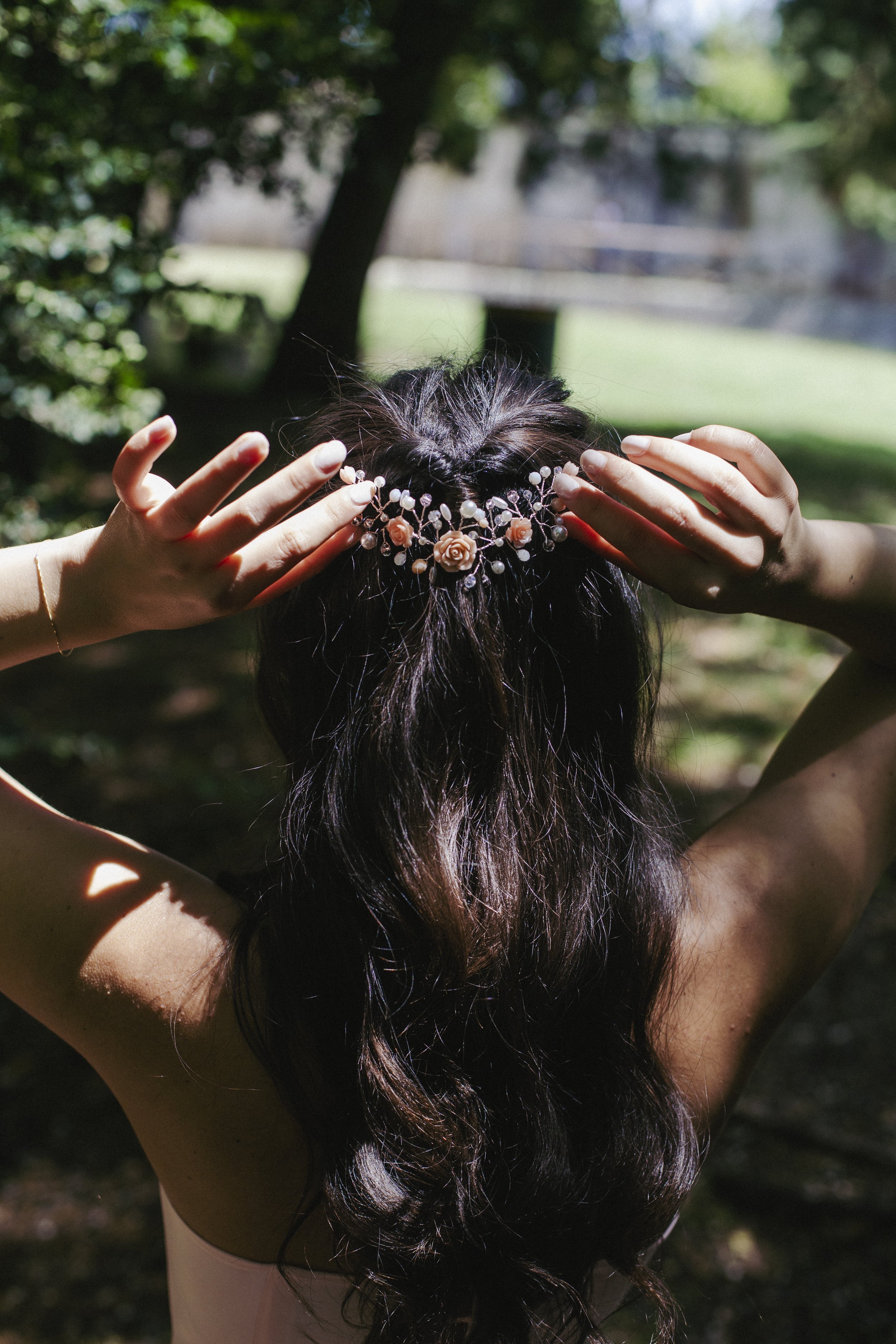Una donna con capelli lunghi e ricci, indossa una corona di fiori rosa e perle, si trova all'aperto tra gli alberi, con luce solare che filtra tra le foglie.