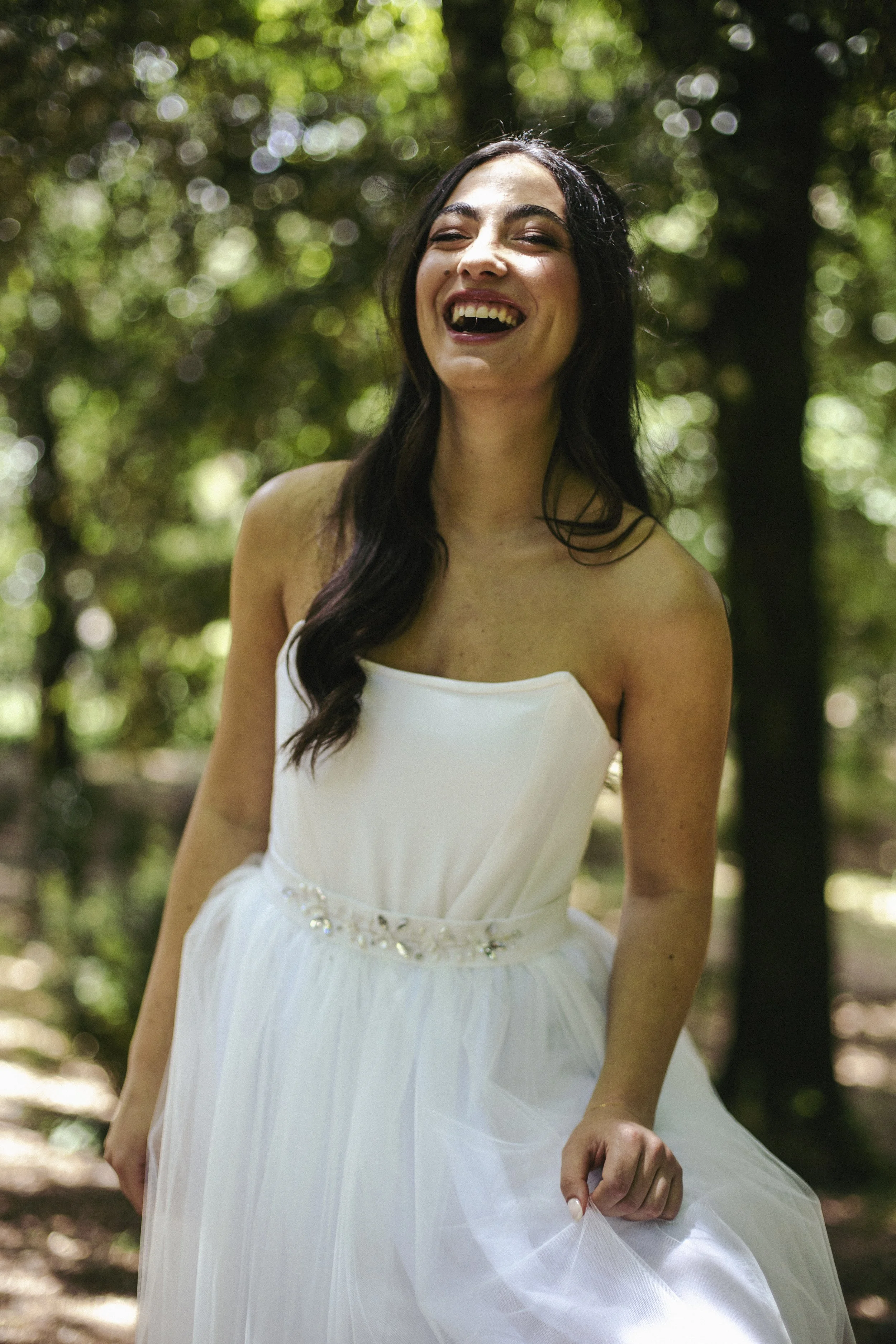 Una donna sorridente con abito bianco in un bosco con luce filtrata tra gli alberi.