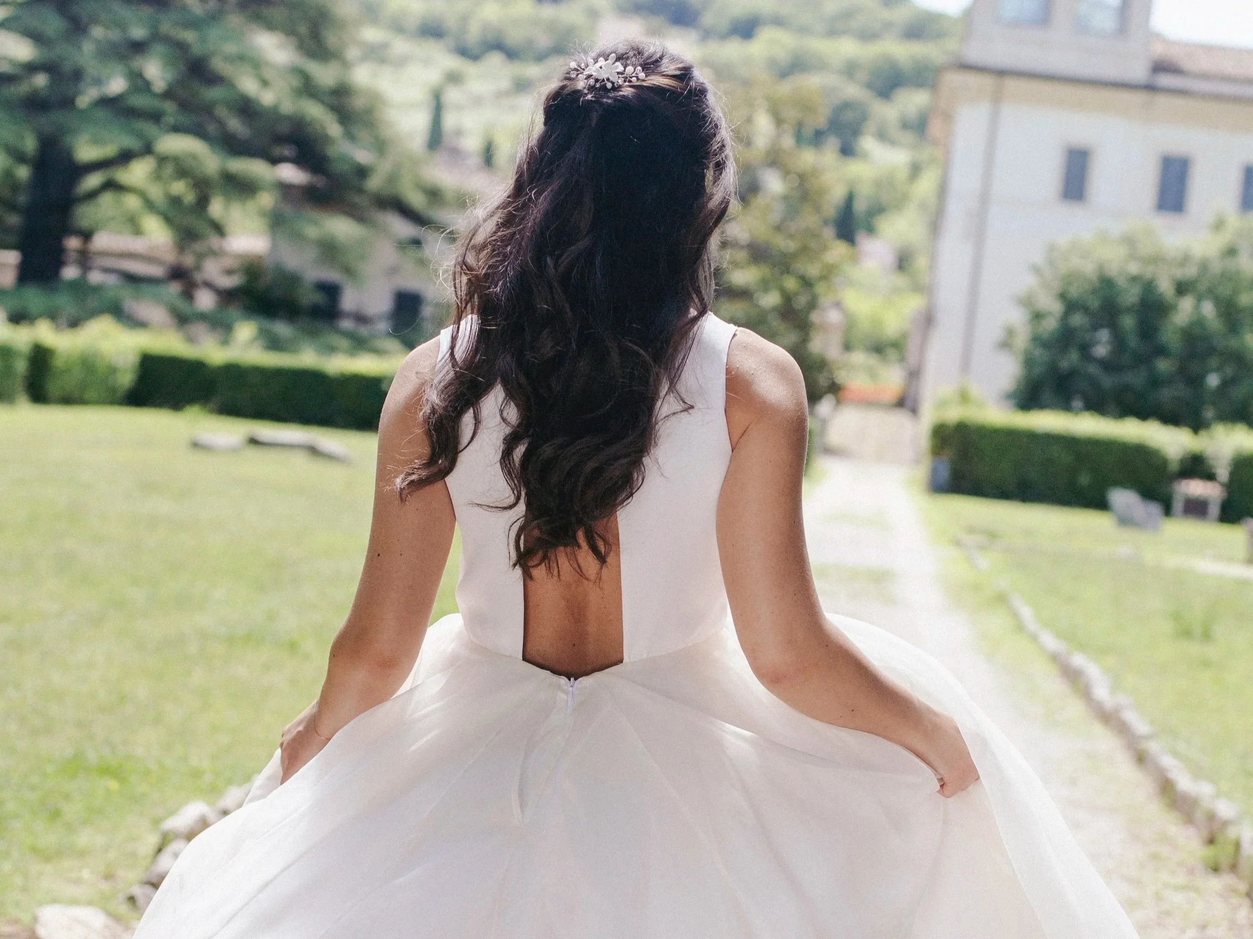 Una donna con capelli lunghi e ricci, indossa un abito da sposa bianco con uno schienale aperto. È seduta all'aperto in un giardino con alberi e un edificio in background, vista la vista del suo vestito che si estende sul prato.