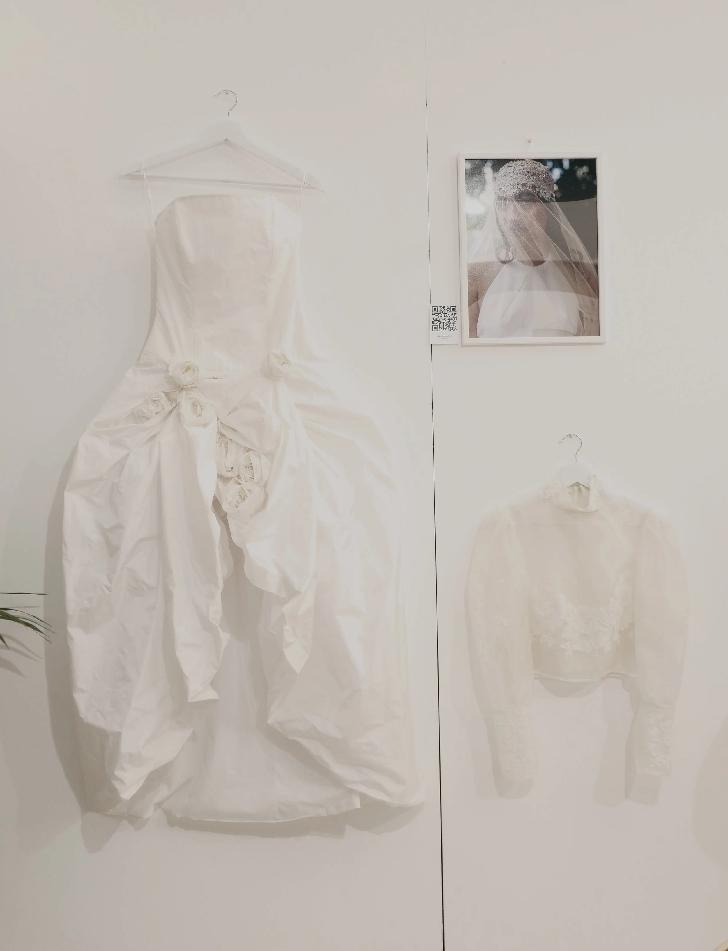 Una parete bianca con un quadro di una sposa, un vestito bianco appeso su una gruccia e alcune rose di carta alle sue basi.