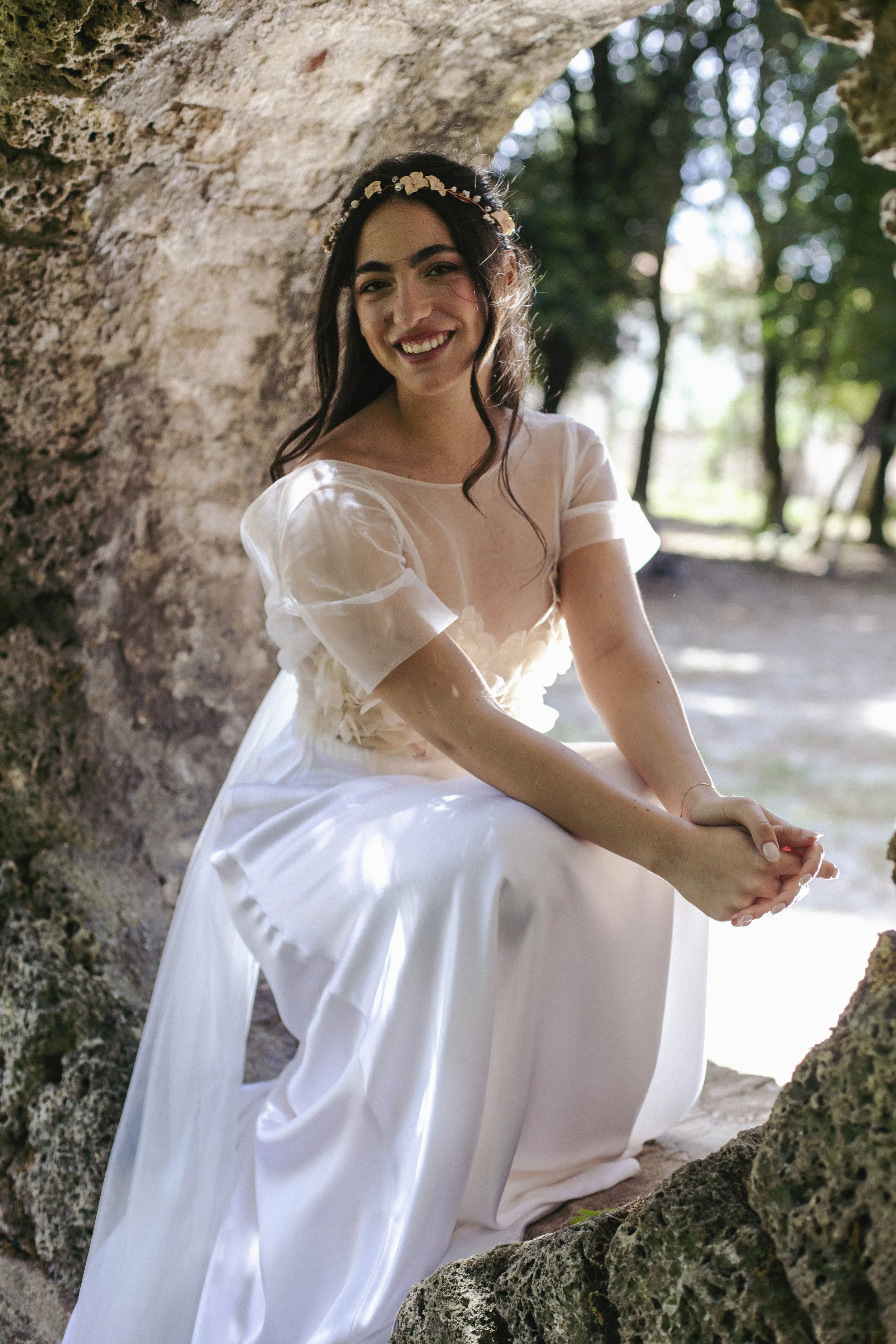 Una donna con un abito bianco, tiara di fiori e trucco leggero, seduta tra rocce e alberi in un ambiente naturale, sorridente