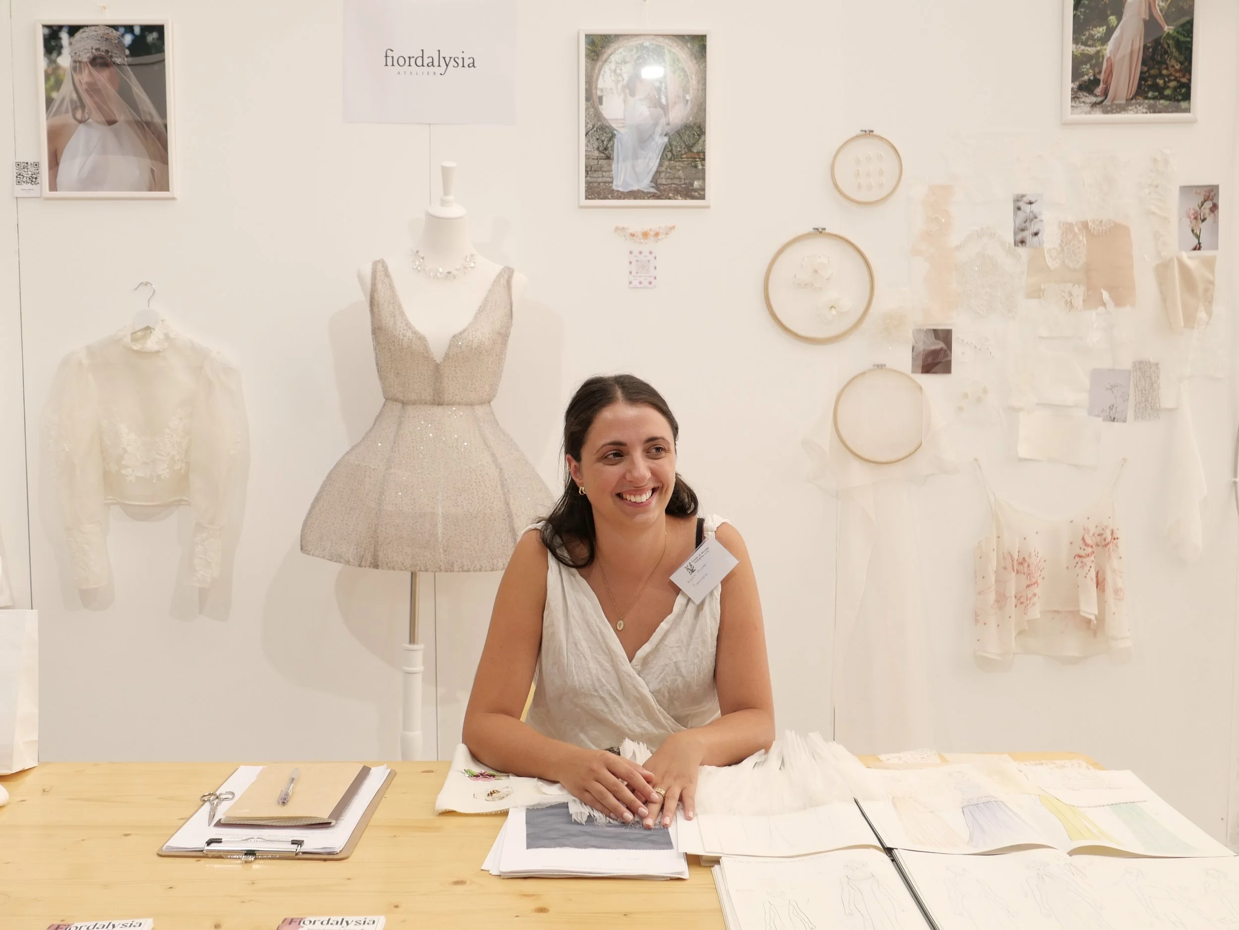 Una donna seduta a un banco di lavoro, circondata da campioni di tessuto, forbici e strumenti di sartoria, con uno sfondo decorato da fotografie e ricami. L'ambiente sembra essere uno studio di moda o atelier di sartoria.