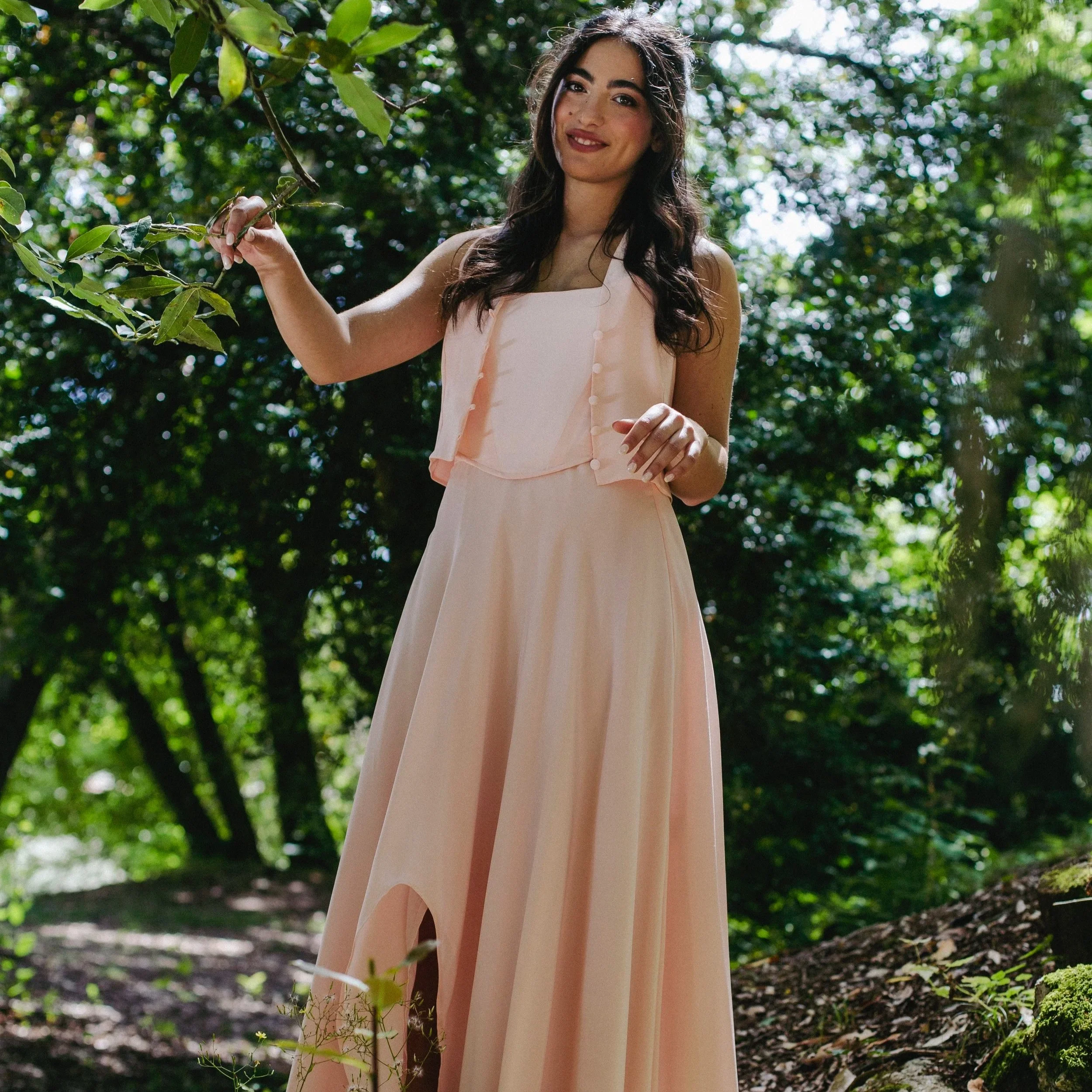 Una giovane donna in abito rosa chiaro si trova in un bosco con foglie verdi e luce naturale che filtra tra gli alberi, mentre si tiene a un ramo di un albero.