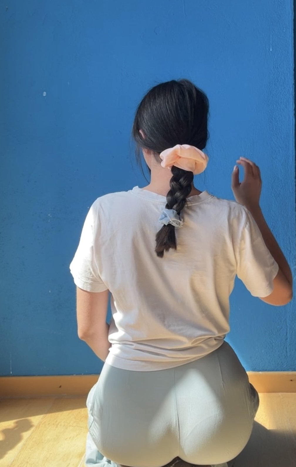 Ragazza con i capelli raccolti in una treccia, poggiata contro una parete blu, vista da dietro.