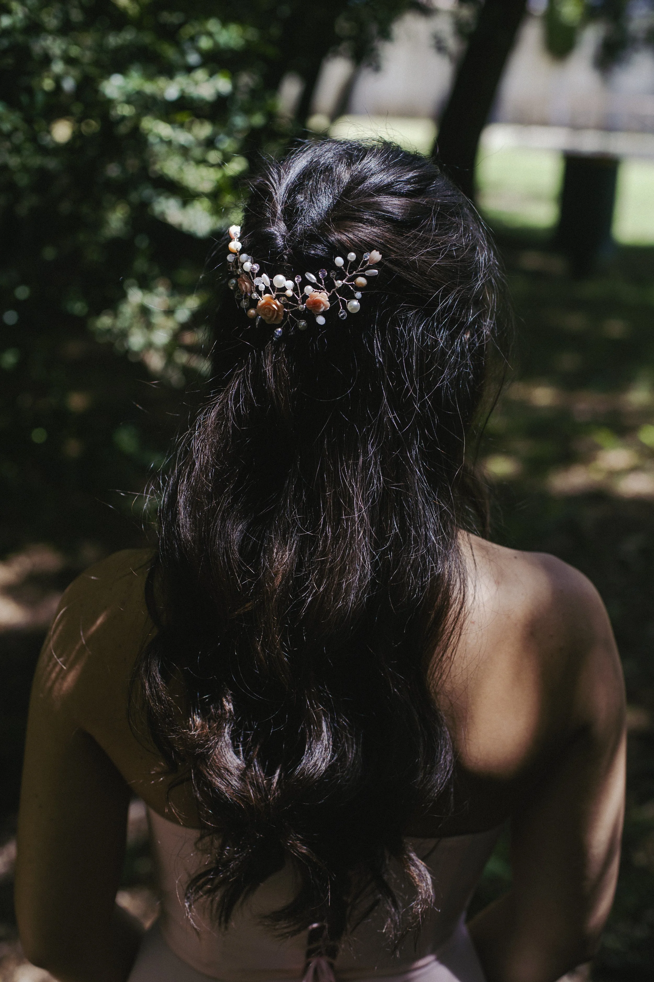 Capelli scuri lunghi e ricci di una donna con un pettine decorato con perle, fiori e perline, in un ambiente naturale con alberi e luce solare.