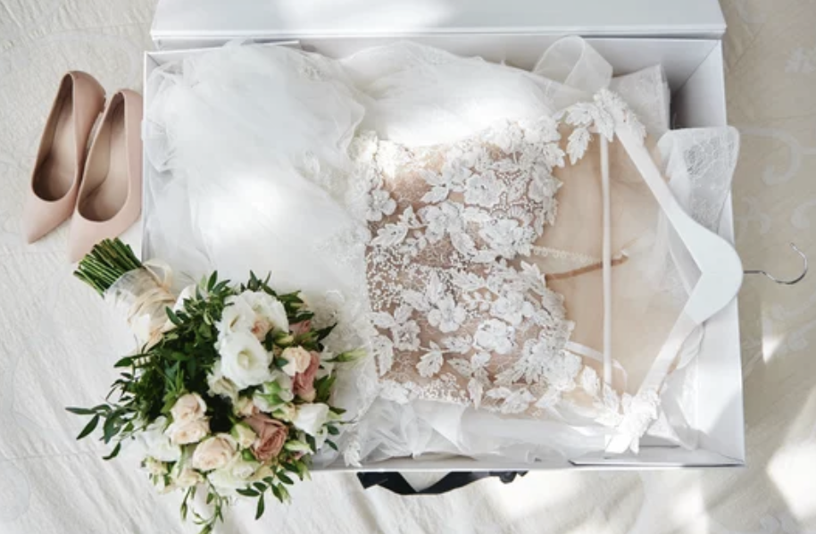 Vestito da sposa di pizzo bianco, bouquet di fiori con rose e verde, paio di scarpe nude, tutto disposto in una scatola.