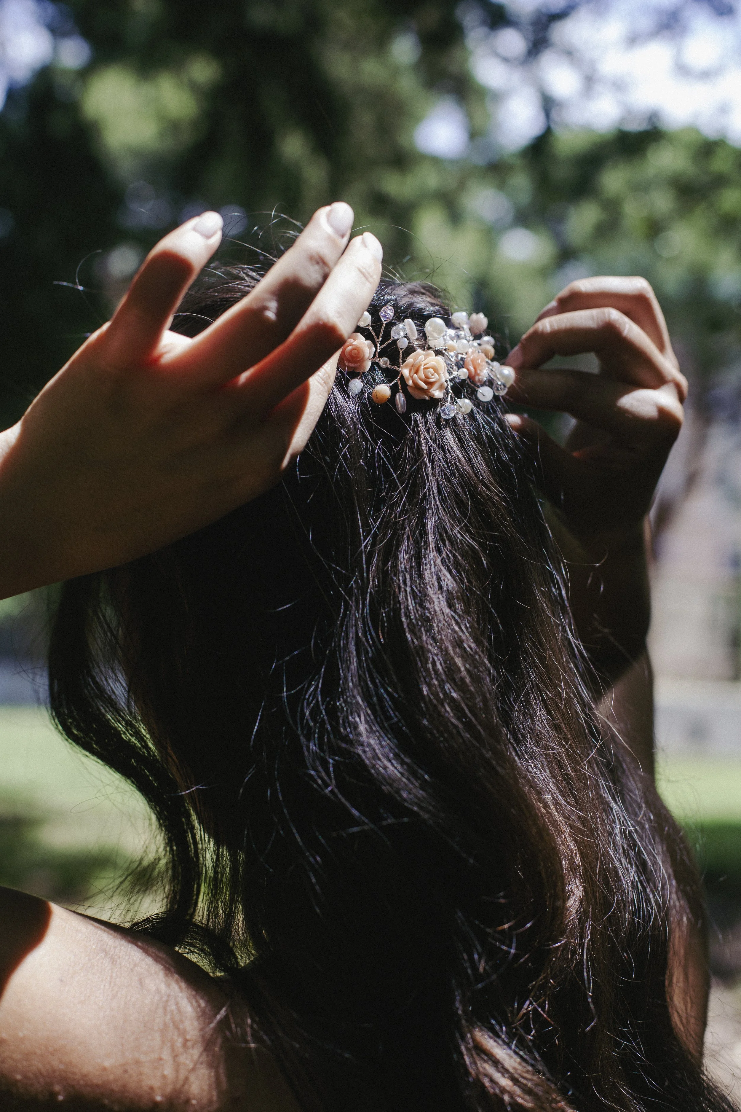 Capelli castani con accessorio di fiori e perle, presenza di mani che sistemano i capelli in un ambiente esterno con luce naturale.
