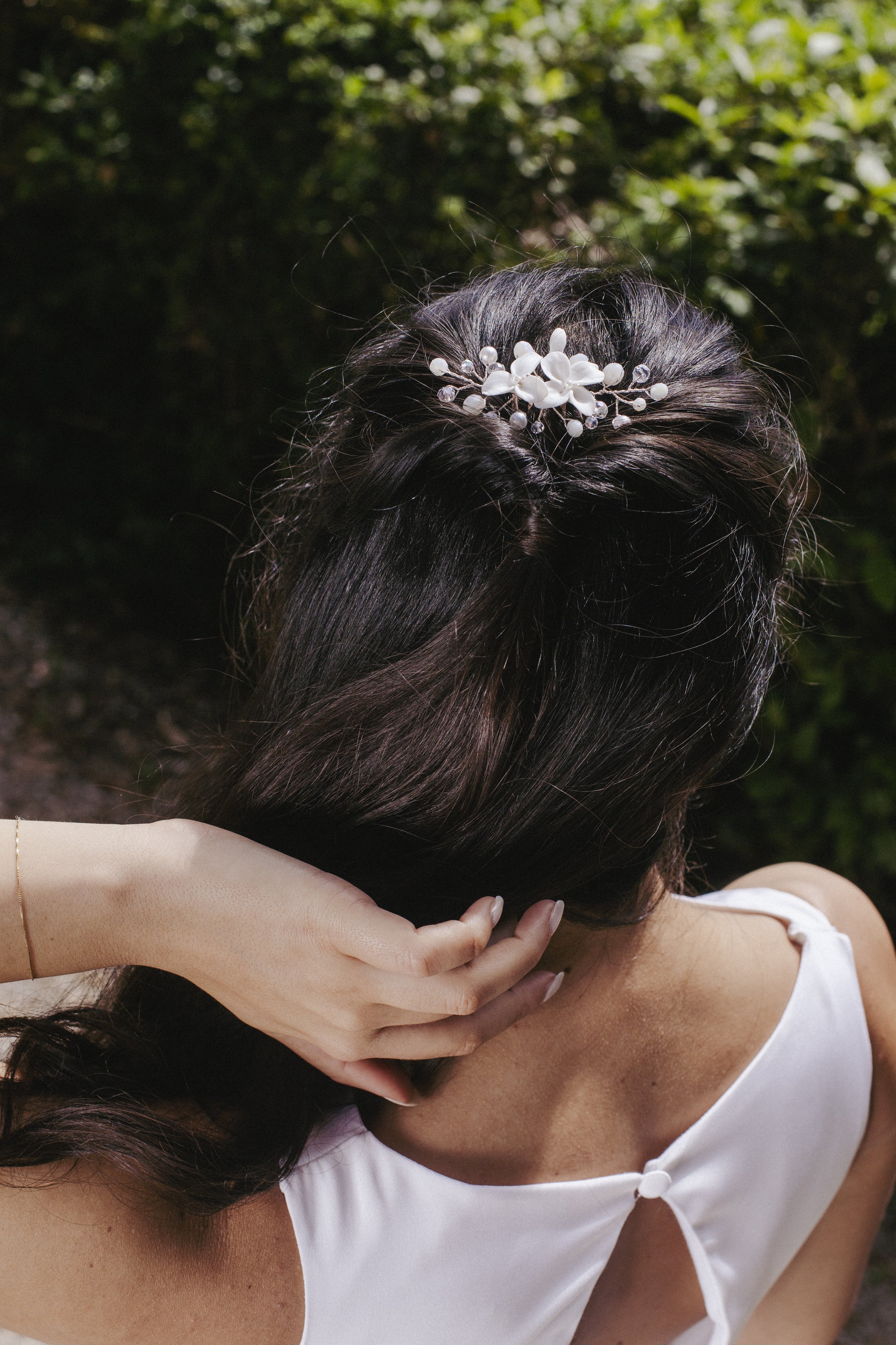 Vista posteriore di una donna con capelli lunghi scuri decorati con un tatuaggio di fiori bianchi e perline, mentre si tiene il collo con una mano, in un ambiente naturale con luci e foglie verdi sullo sfondo.