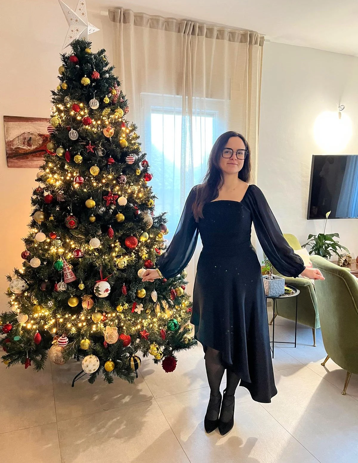 Una donna con abito nero elegante e occhiali neri sta davanti a un albero di Natale decorato con luci e ornamenti rossi, dorati e altri colori, in una stanza con tende chiare e un televisore a parete.