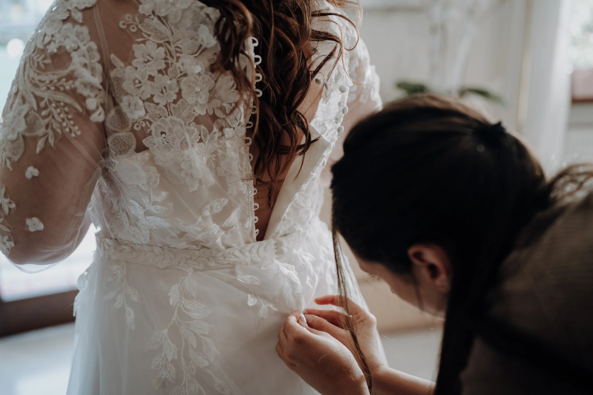 Una donna con un abito da sposa bianco ricamato riceve aiuto da un'altra persona, forse mentre si prepara per un matrimonio.