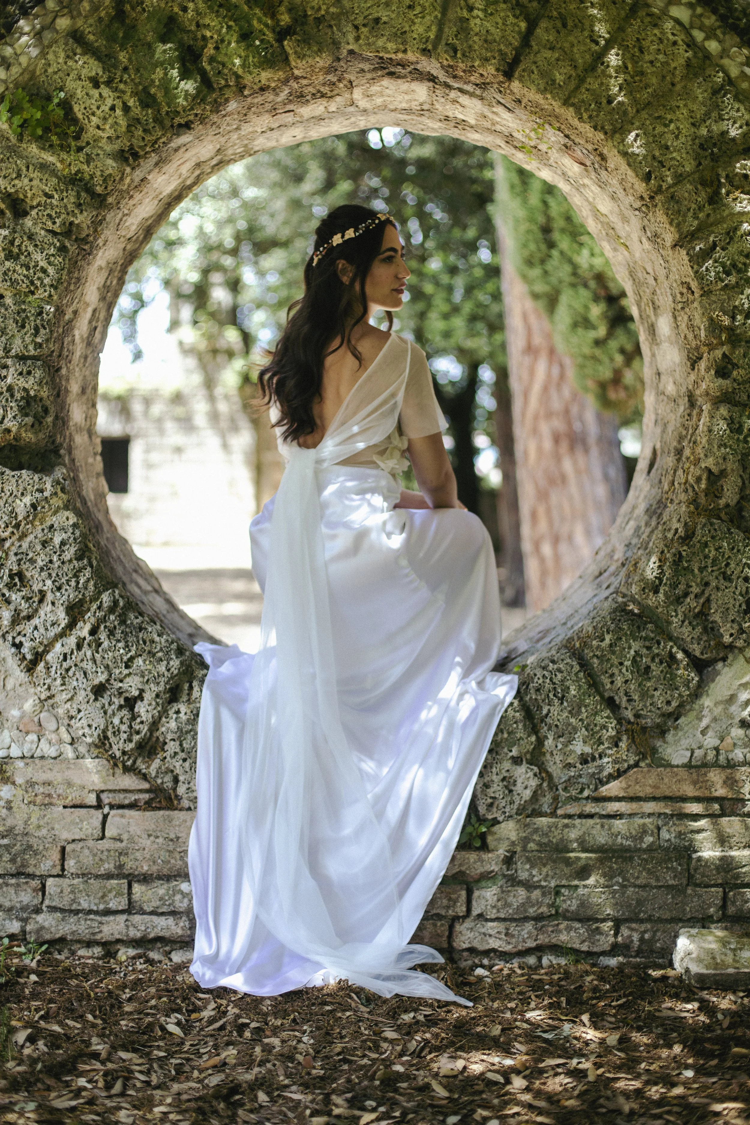 Una donna indossa un abito bianco con un fiore tra i capelli, seduta in un anello di pietra in un ambiente naturale circondato da alberi e foglie.