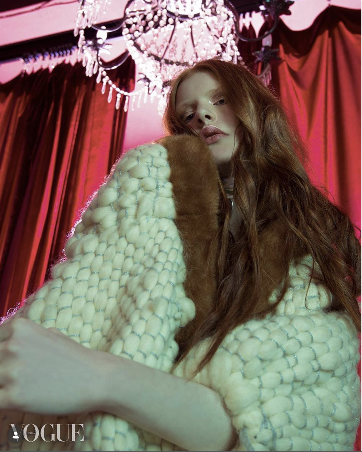 Ragazza con lunghi capelli ricci rossi indossa un cappotto beige in stile peluche e si trova in una stanza con tende rosse e un lampadario di cristallo.