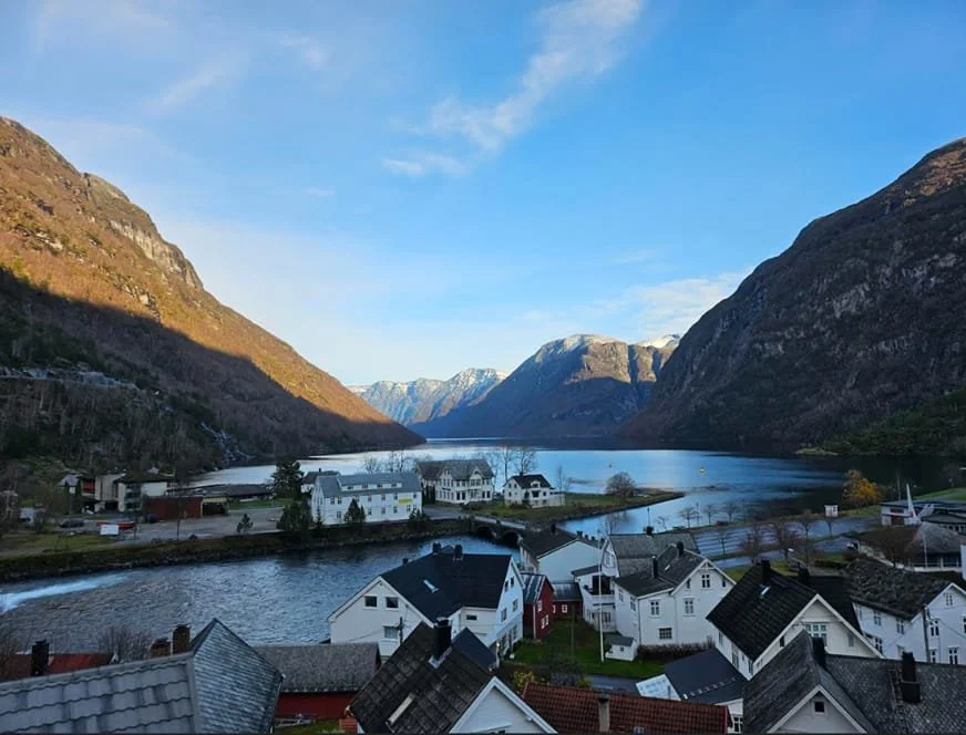 Smal landsby ved en fjord omgitt av fjell med snødekte topper, solfylt dag.
