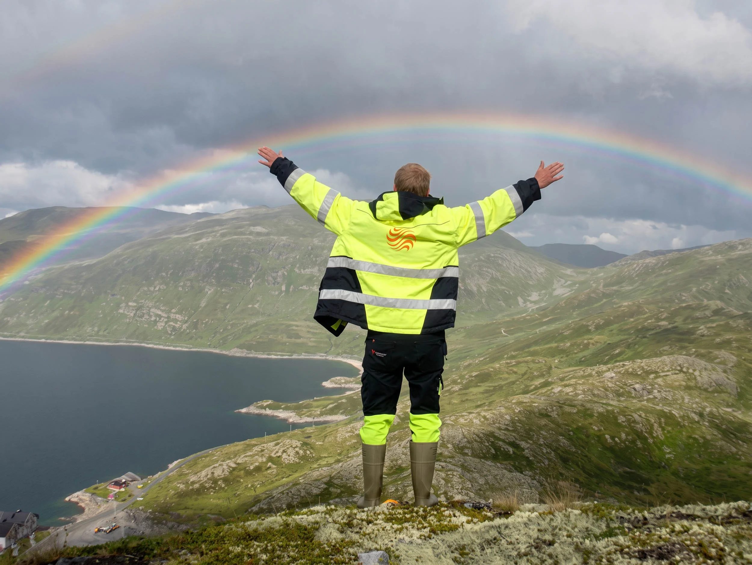 Person i gul refleksjakke med armene utstrakt foran en regnbue over et fjellandskap og en innsjø.