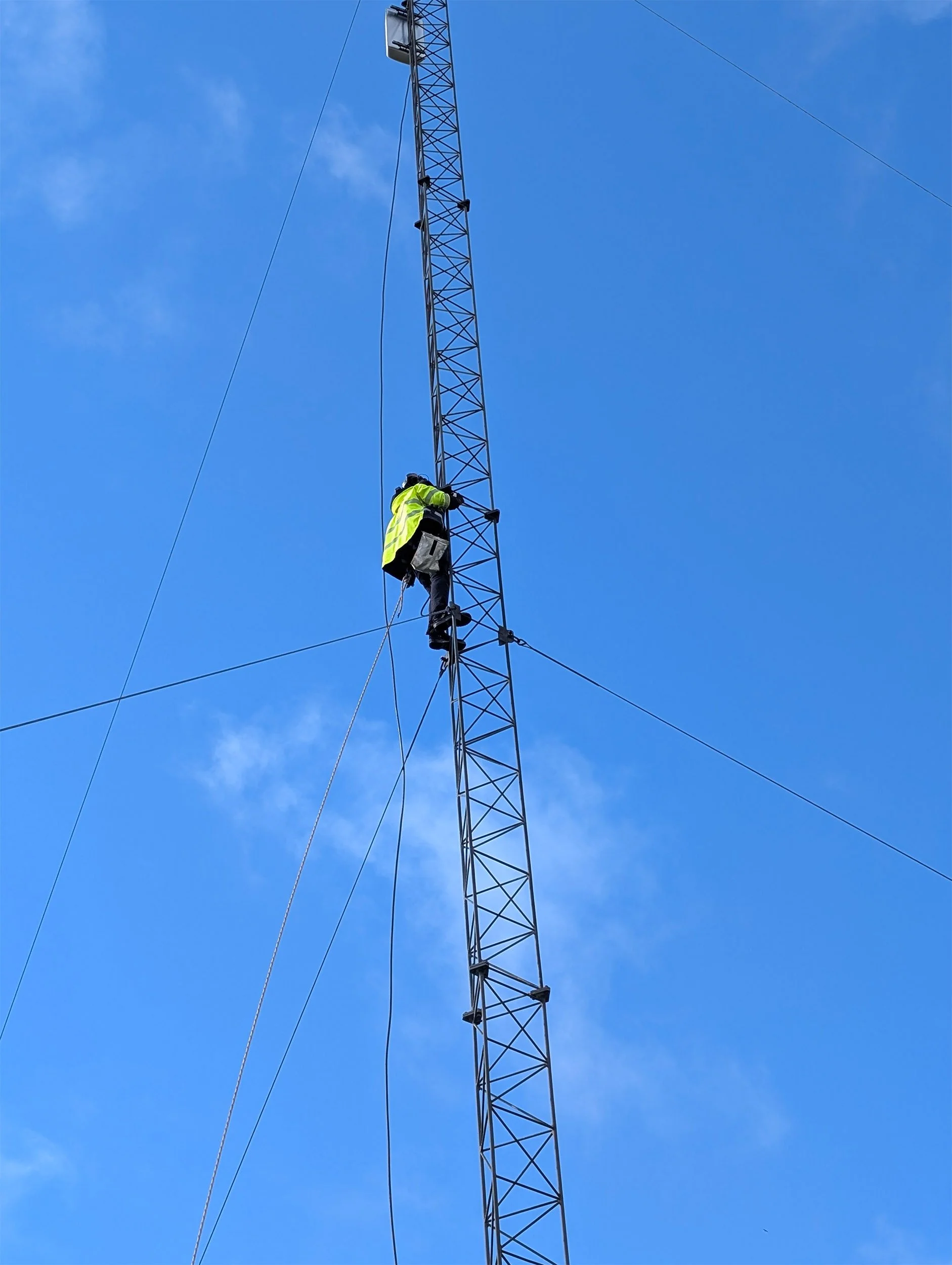 Arbeider i verneutstyr klatrer i en høy gittermast mot blå himmel.