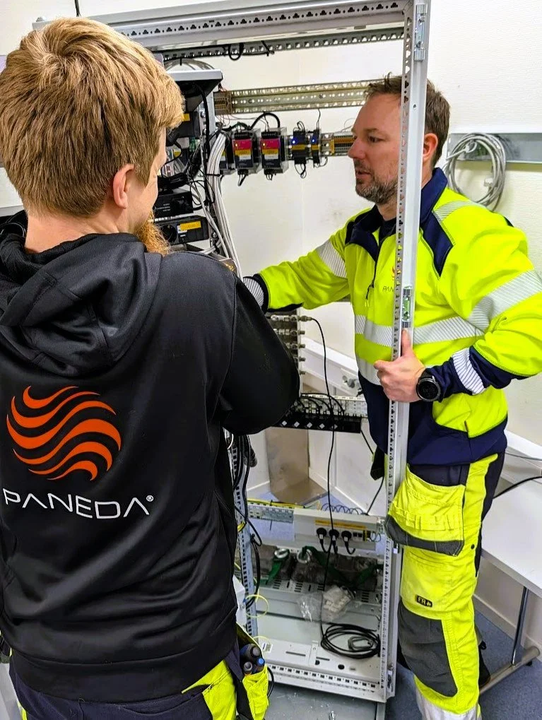 To teknikere arbeider med elektrisk utstyr i et teknisk skap; én har på seg Paneda-jakke og den andre bruker fluorescerende verneklær.
