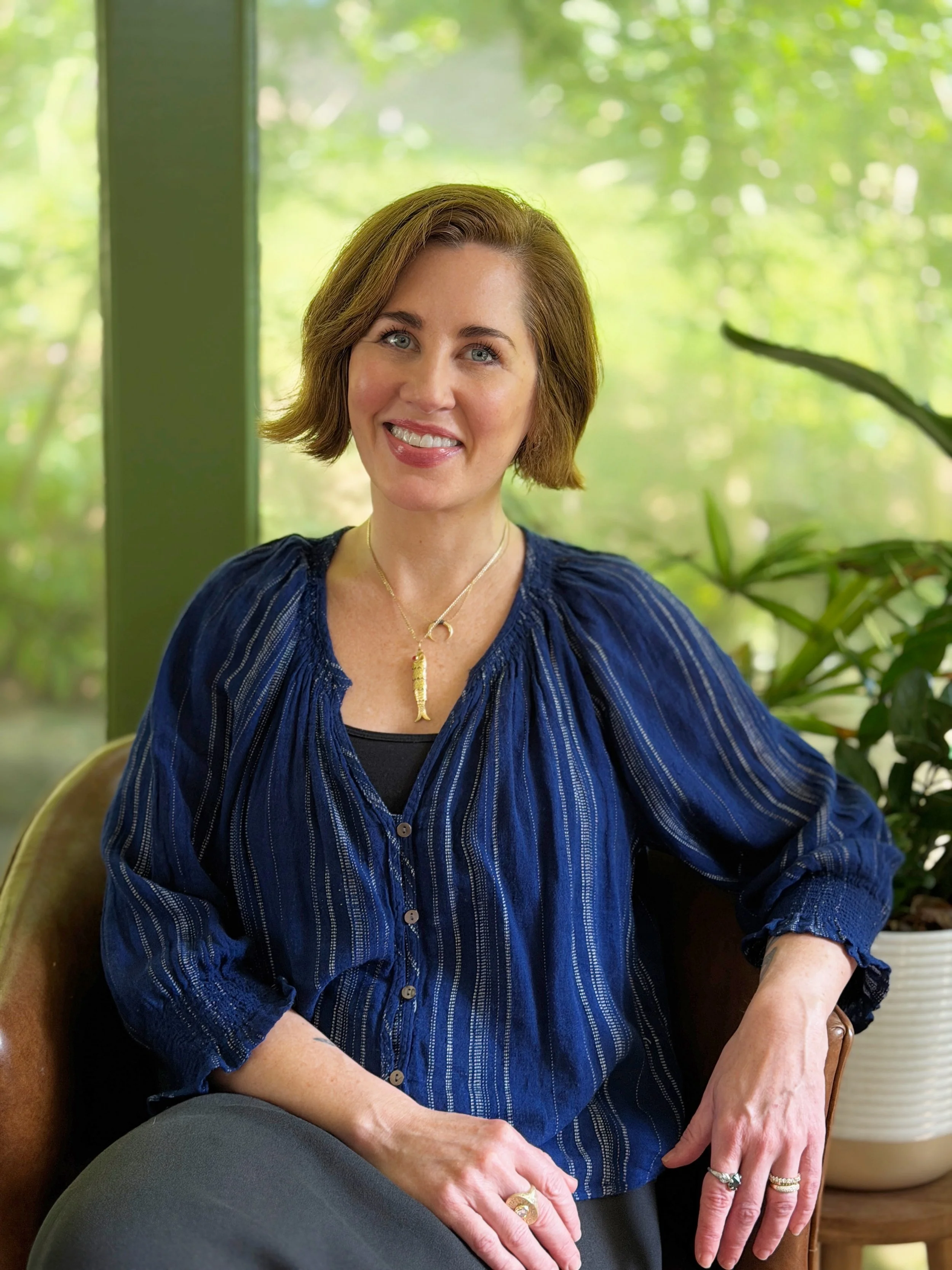 A woman with short brown hair and blue eyes, smiling while sitting indoors with a green, blurred outdoor background, wearing a blue striped blouse and rings on her fingers.