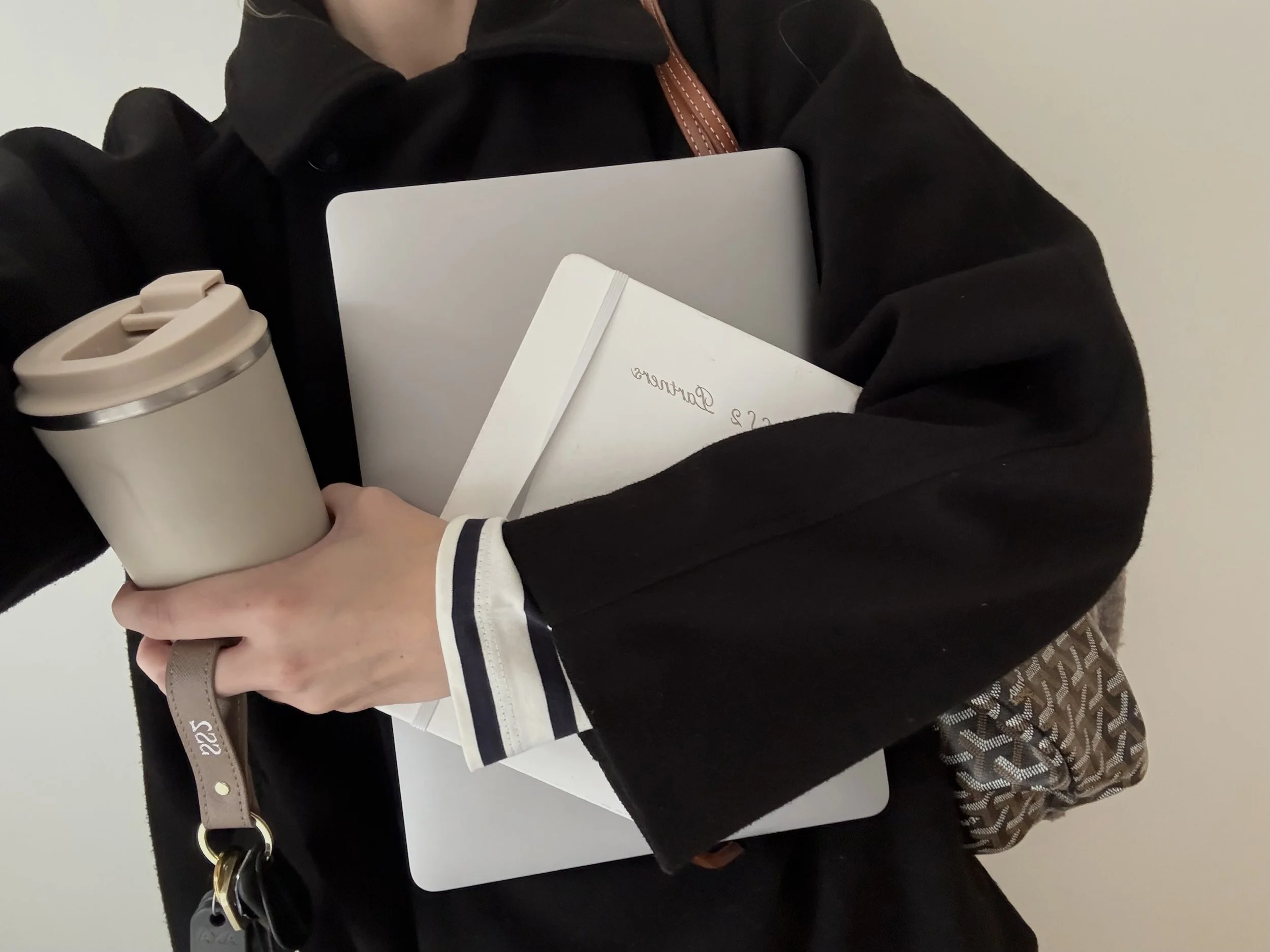 Person holder en computer, en notesbog, en termokop, nøgler og en taske. Personen er iført sort jakke med hvide kanter på ærmerne.
